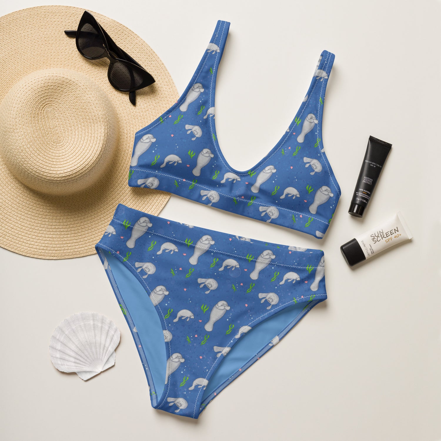 High-waisted bikini with hand-illustrated manatee pattern on a blue background. Made from recycled polyester combined with stretchable fabric. Has double layers and removable pads. Flat lay view shown with sun hat, sunglasses, sunscreen and seashell.