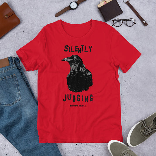 Unisex red colored t-shirt. Features vertical image of silently judging text above and below black crow wearing a monocle.  Shown on ground by pants, shoes, glasses, wallet, and watch.
