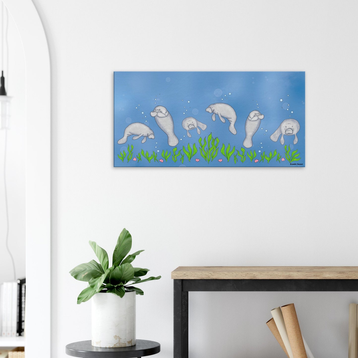 20 by 40 inch slim canvas wall art print featuring cute illustrated manatees swimming above the seabed. Shown on wall above wooden end table and potted plant on stand.