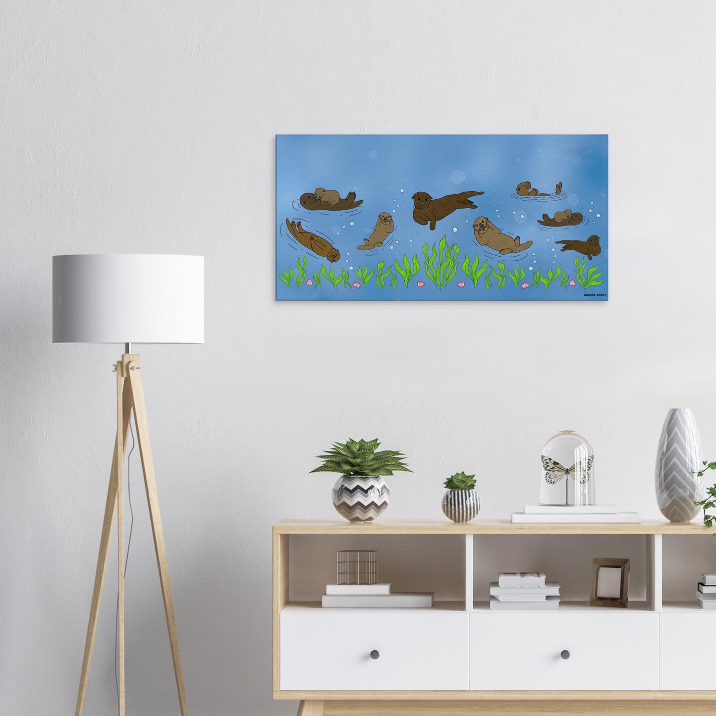 20 by 40 inch slim canvas wall art print of sea otters swimming along the seabed. Shown on wall above wooden end table and potted plants and next to a lamp.