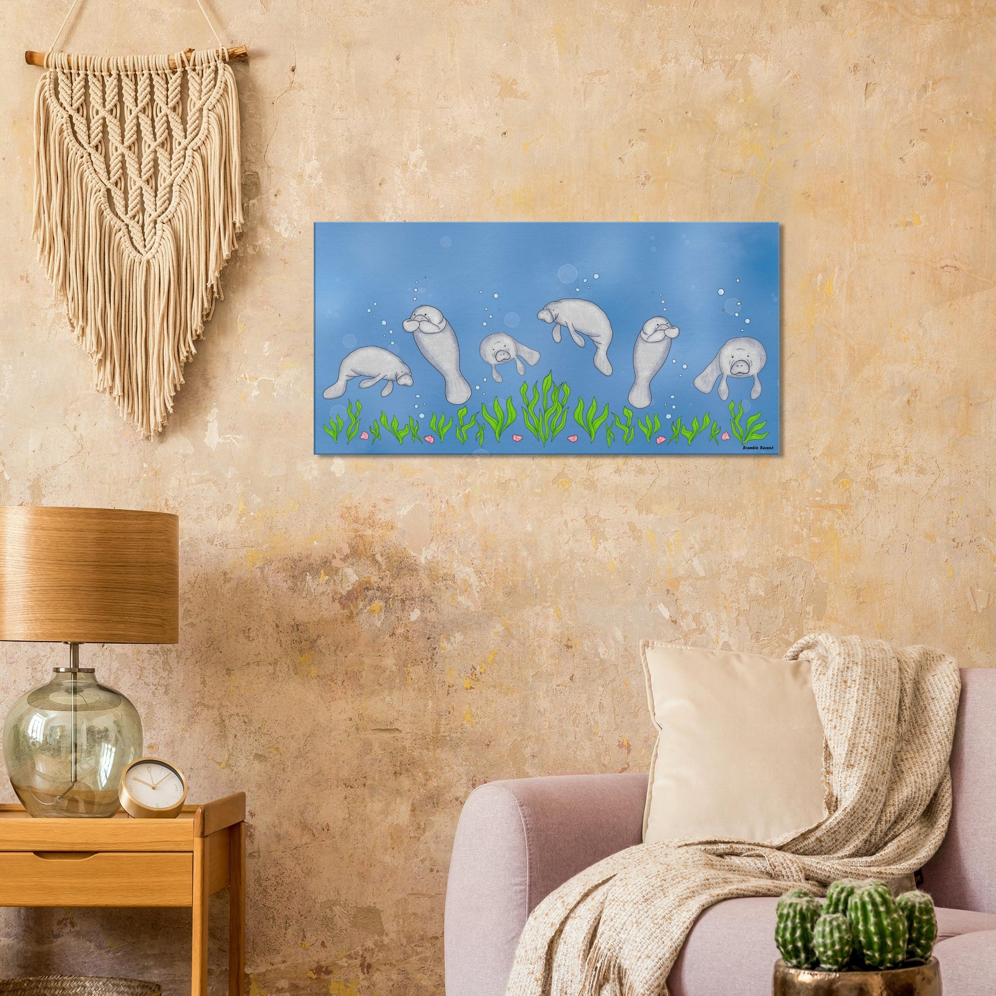 20 by 40 inch slim canvas wall art print featuring cute illustrated manatees swimming above the seabed. Shown on  beige wall by macrame, lamp, wooden end table, and pink sofa.