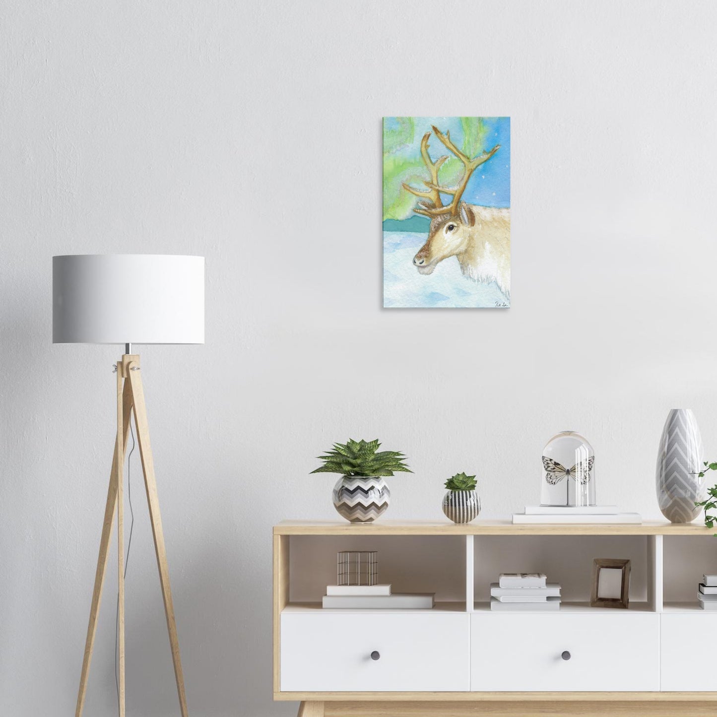 a44a98c2-f6f8-45b4-b1aa-537c697f4ab216 by 24 inch slim canvas print of Heather Silver's watercolor painting, northern lights reindeer. Shown on wall above shelves with potted plants and a lamp.