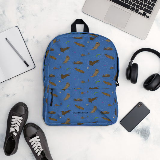 Medium-sized backpack featuring sea otters, seashells, seaweed and sea urchins on a blue background. Made with water resistant polyester, ergonomic straps, and comfort mesh back. Has a compartment that can hold a 15 inch laptop. Has a hidden zipper pocket on the back. Shown by laptop, headphones, shoes, phone, and notebook.