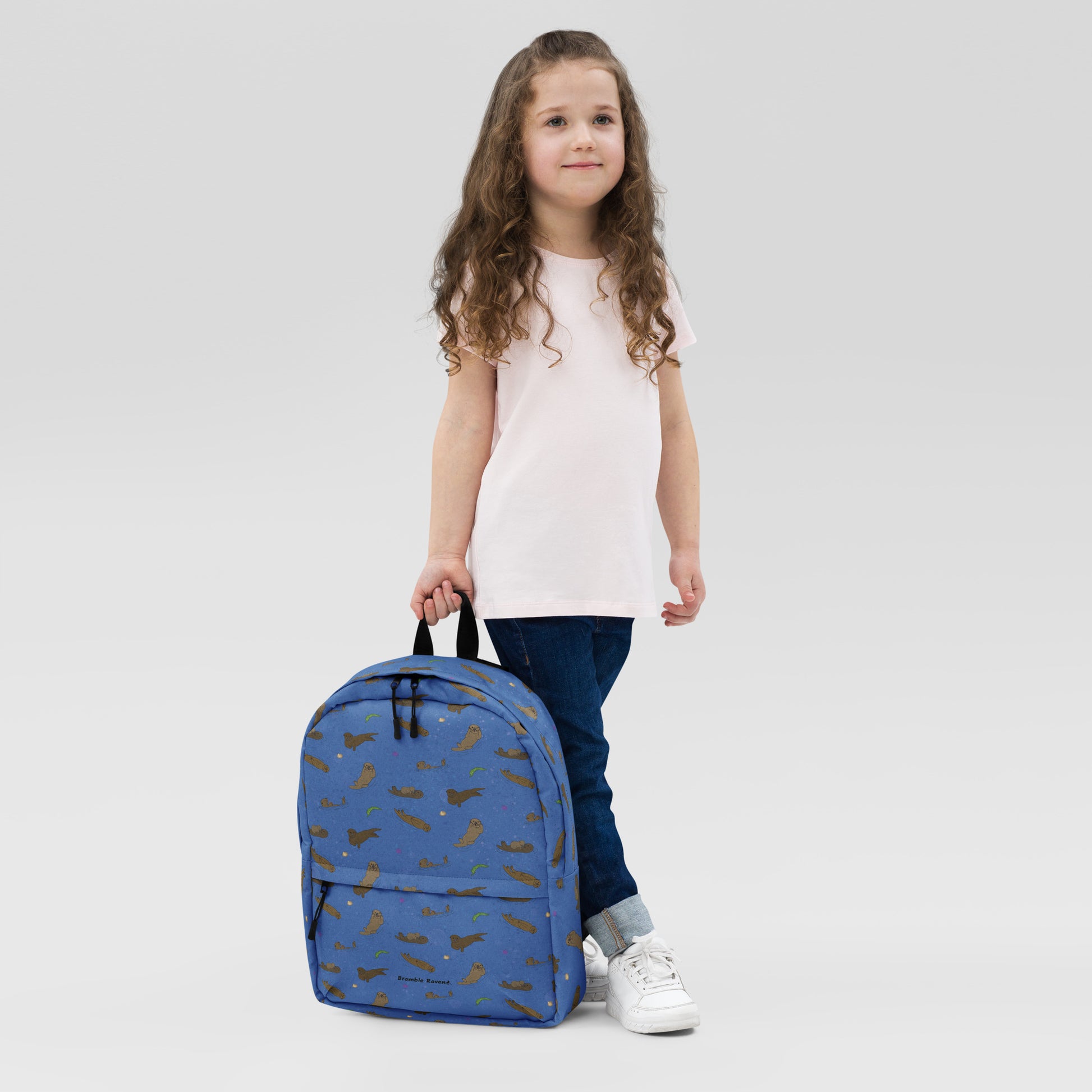 Medium-sized backpack featuring sea otters, seashells, seaweed and sea urchins on a blue background. Made with water resistant polyester, ergonomic straps, and comfort mesh back. Has a compartment that can hold a 15 inch laptop. Has a hidden zipper pocket on the back. Shown being held by a girl.