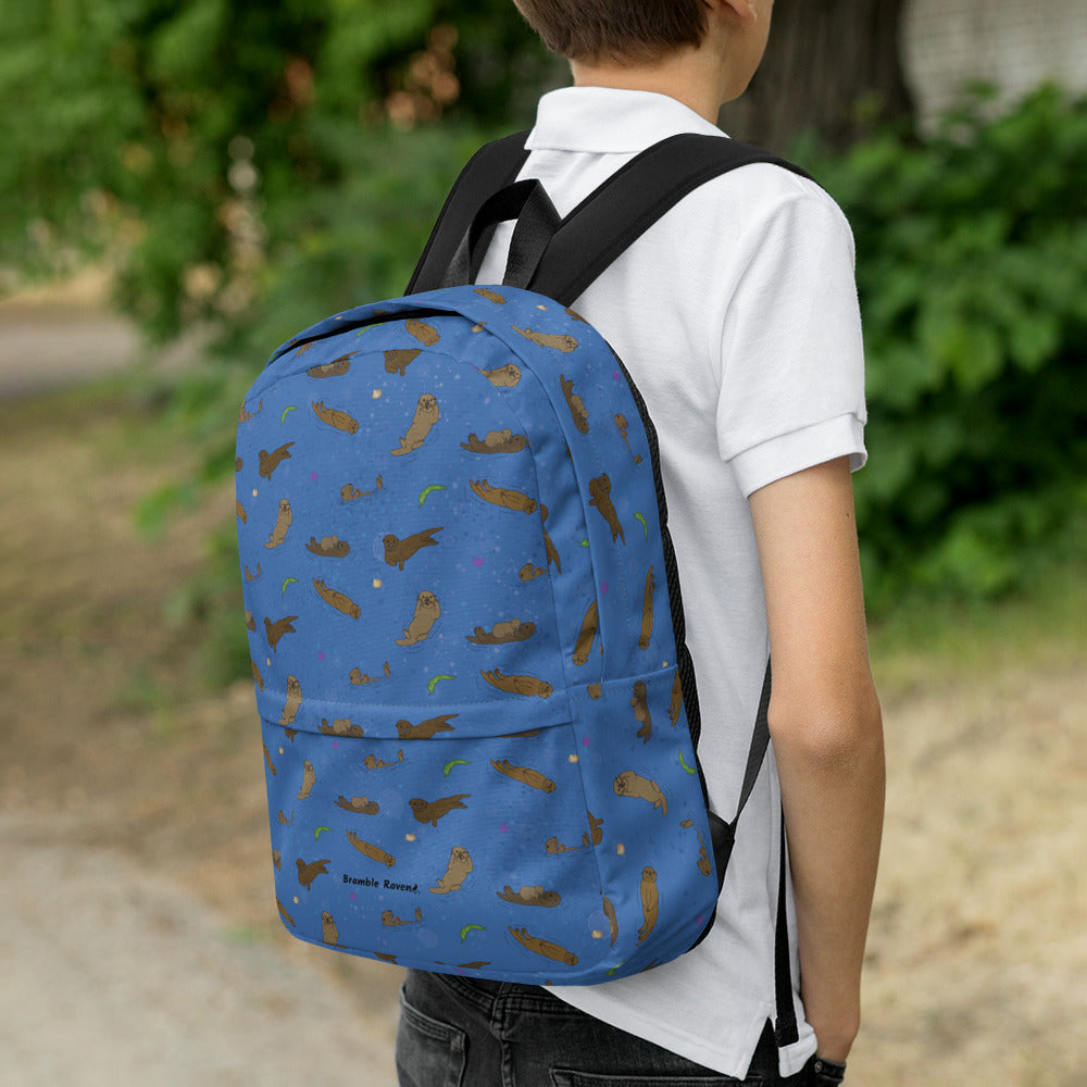 Medium-sized backpack featuring sea otters, seashells, seaweed and sea urchins on a blue background. Made with water resistant polyester, ergonomic straps, and comfort mesh back. Has a compartment that can hold a 15 inch laptop. Has a hidden zipper pocket on the back. Shown on the back of a boy.
