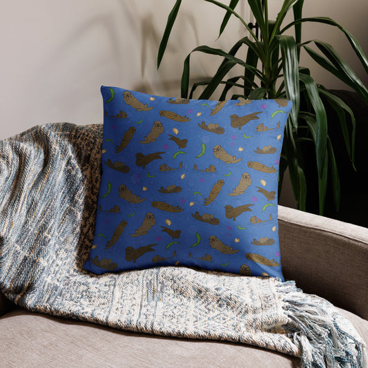 22 by 22 inch accent pillow. Double-sided featuring original hand illustrated sea otters with urchins, seaweed, and shells on an ocean blue background. Shown on a tapestry blanket on a tan sofa.