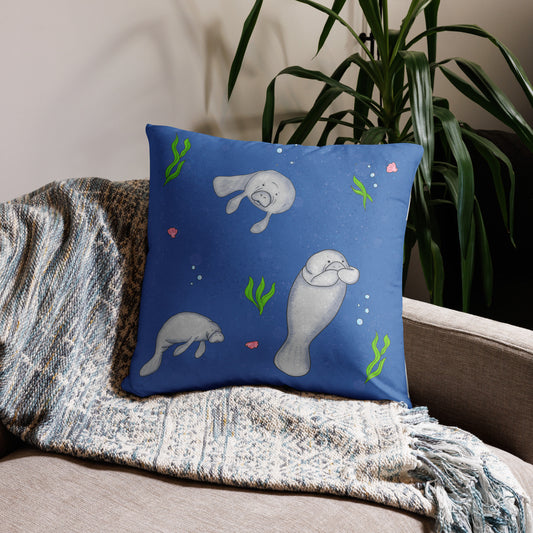 22 by 22 inch accent pillow. Features hand illustrated manatees, surrounded by seaweed, seashells, and bubbles on an ocean blue background. Large design on front, small patterned design on back. Shown on throw blanket on a tan sofa with a potted plant behind.