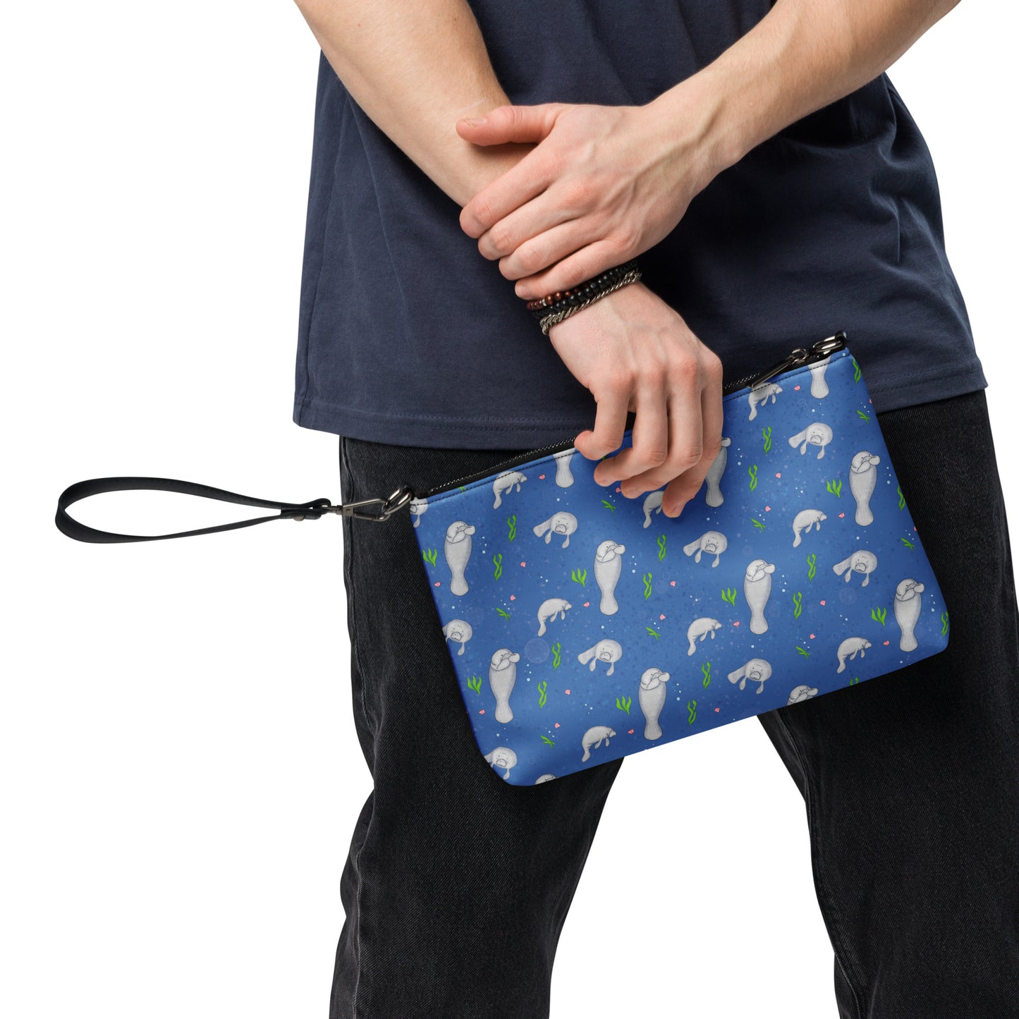 Manatee crossbody bag. Faux leather with polyester lining and dark grey hardware. Comes with adjustable removable wrist and shoulder straps. Back view of bag with manatees patterned on a dark blue background. Shown in model's hand.