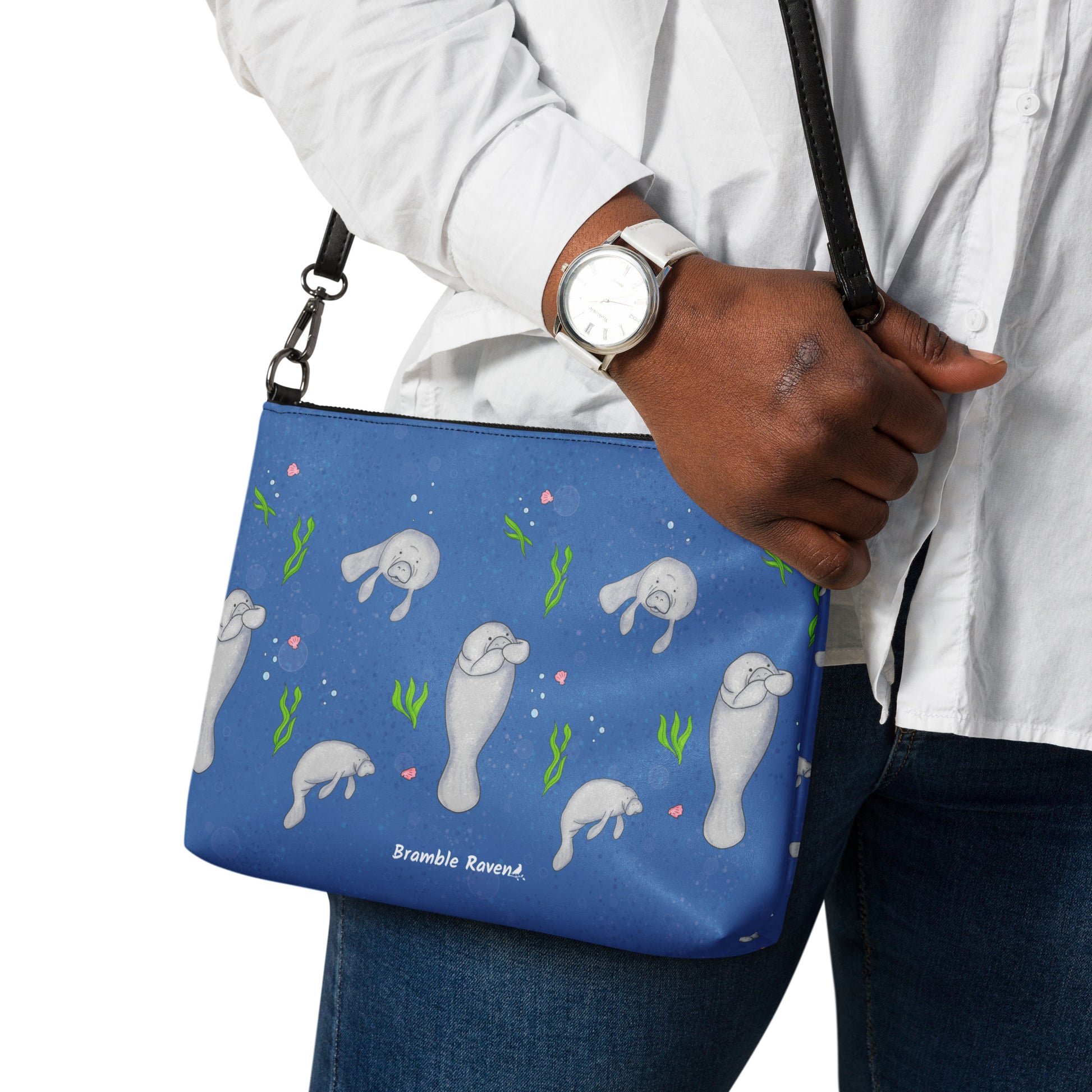 Manatee crossbody bag. Faux leather with polyester lining and dark grey hardware. Comes with adjustable removable wrist and shoulder straps. Front view of bag with manatees on a dark blue background. Shown on shoulder of model.
