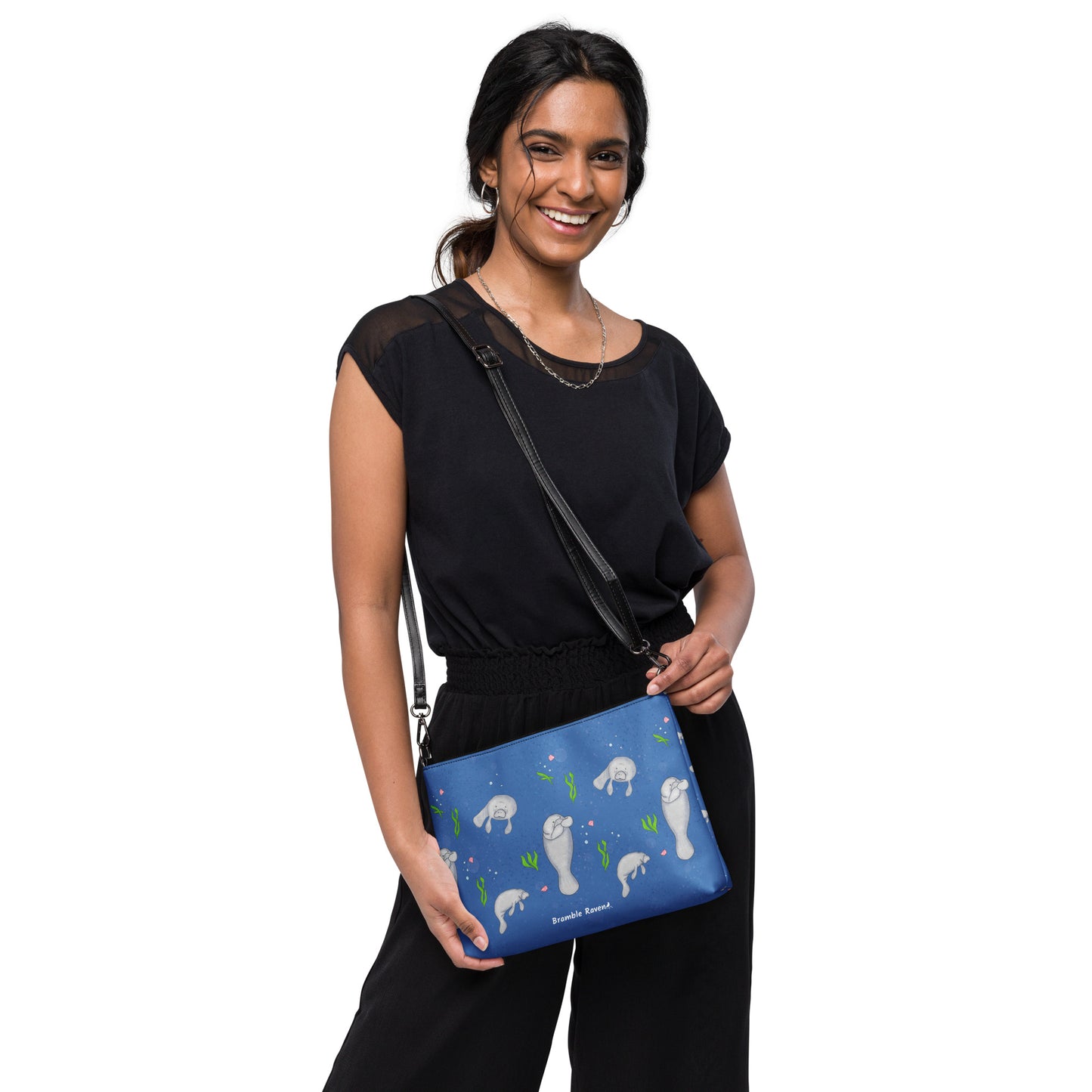 Manatee crossbody bag. Faux leather with polyester lining and dark grey hardware. Comes with adjustable removable wrist and shoulder straps. Front view of bag with manatees on a dark blue background. Shown on shoulder of female model.