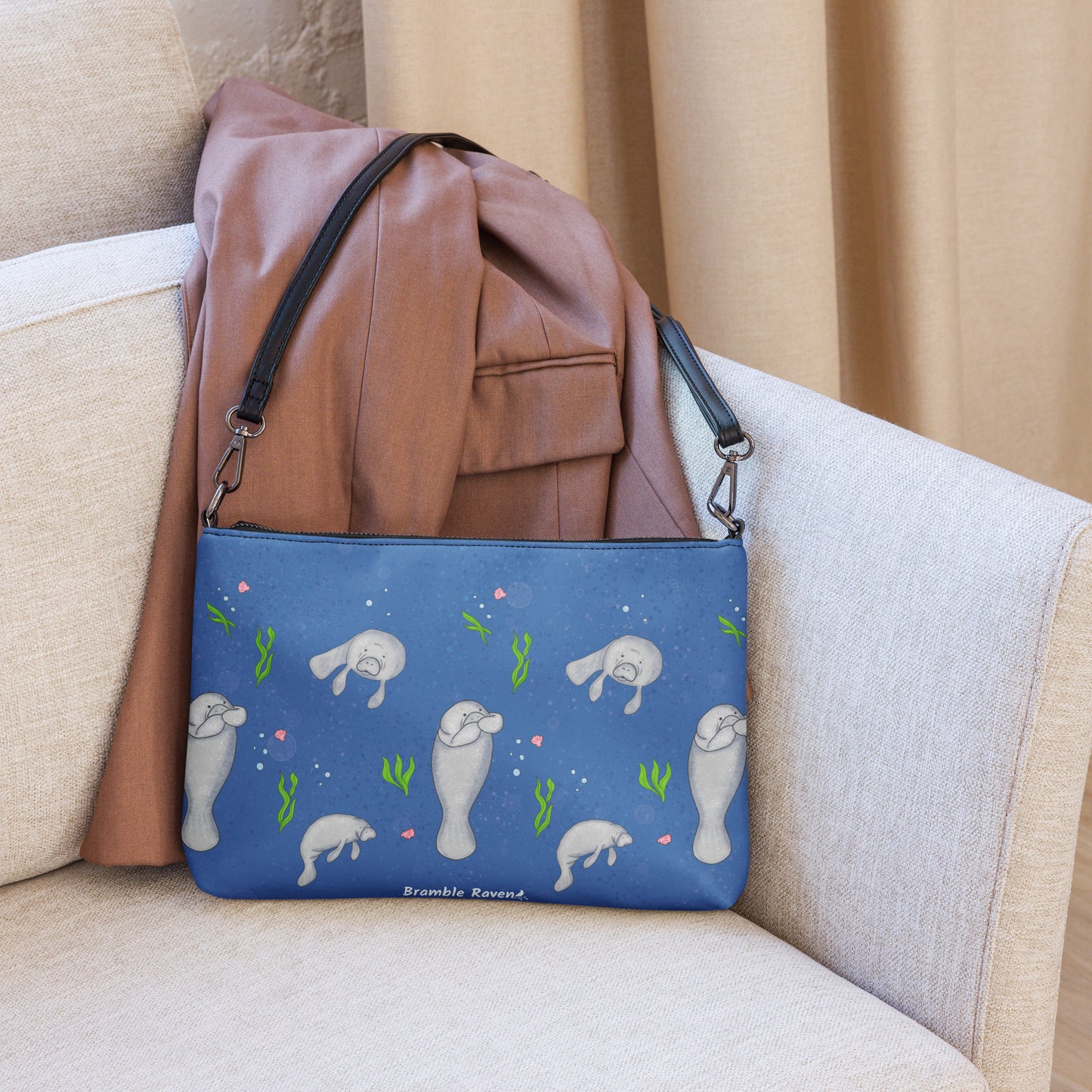 Manatee crossbody bag. Faux leather with polyester lining and dark grey hardware. Comes with adjustable removable wrist and shoulder straps. Front view of bag with manatees on a dark blue background. Shown on couch by leather jacket.