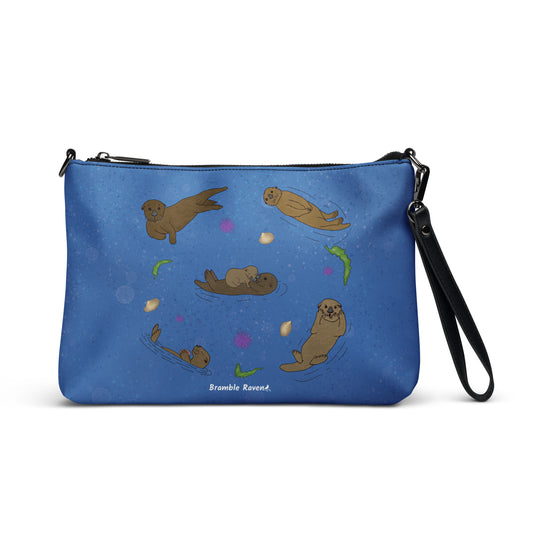 Sea Otter crossbody bag. Faux leather with polyester lining and dark grey hardware. Comes with adjustable removable wrist and shoulder straps. Front view of bag with a circle of sea otters on a dark blue background.
