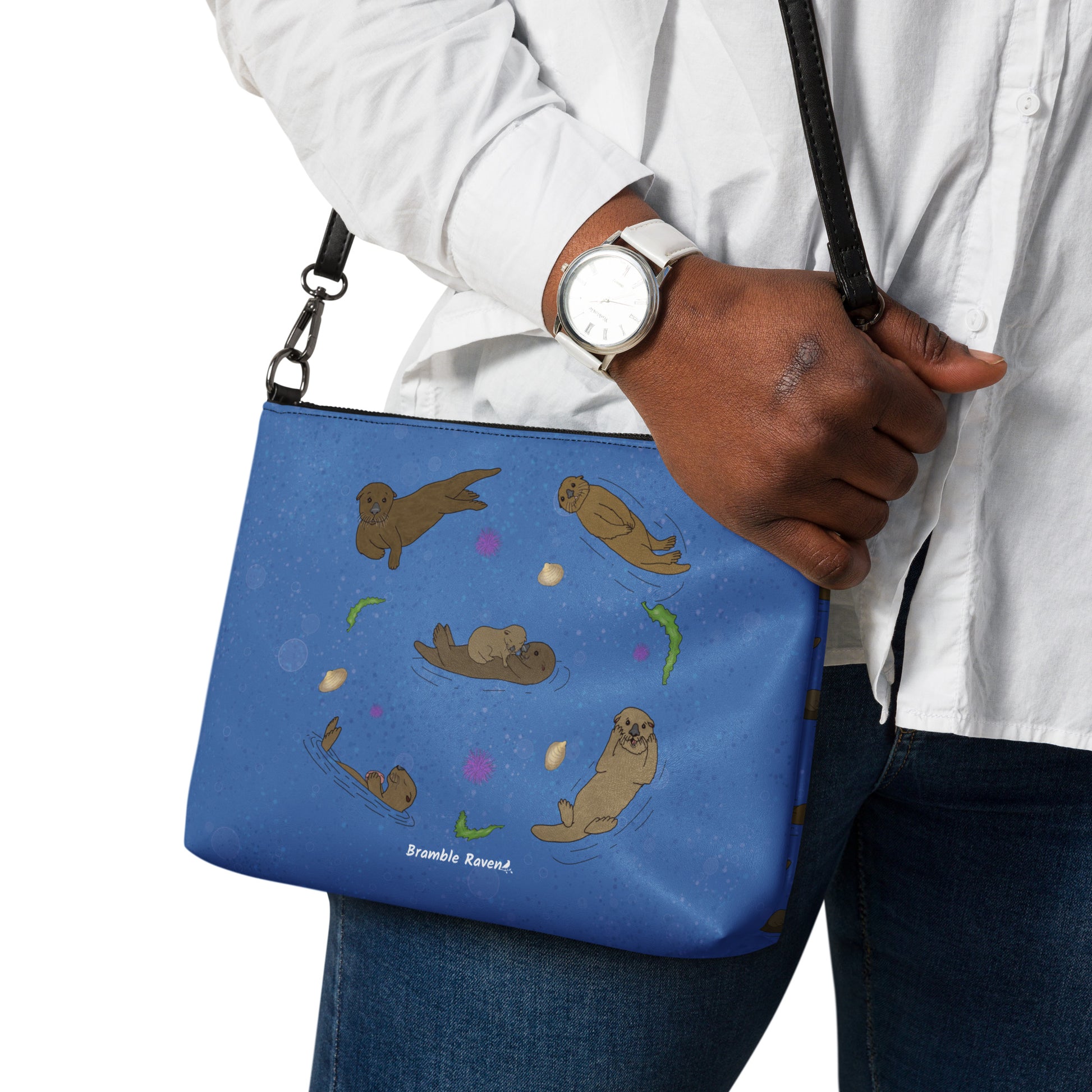 Sea Otter crossbody bag. Faux leather with polyester lining and dark grey hardware. Comes with adjustable removable wrist and shoulder straps. Front view of bag with a circle of sea otters on a dark blue background. Shown on model's shoulder.