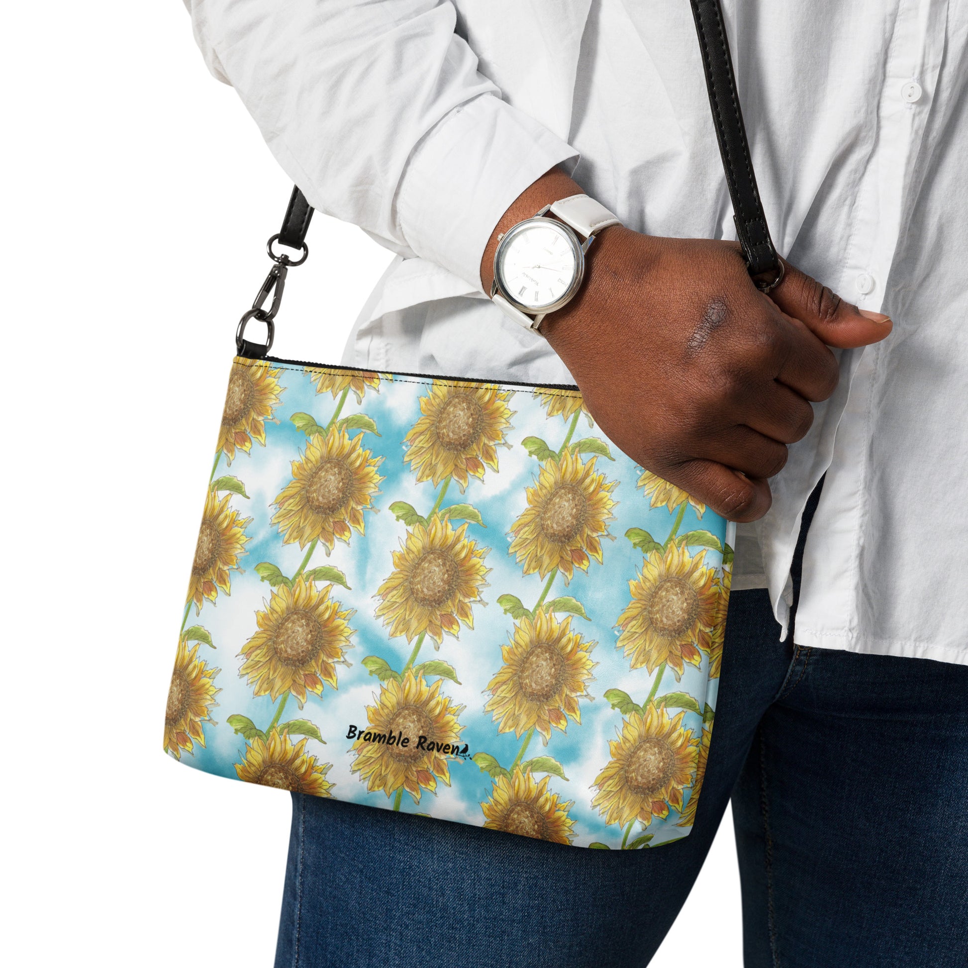Sunflower crossbody bag. Faux leather with polyester lining and dark grey hardware. Comes with adjustable removable black wrist and shoulder straps. Features patterned design of watercolor sunflowers against a cloudy blue background. Shown on model's shoulder.