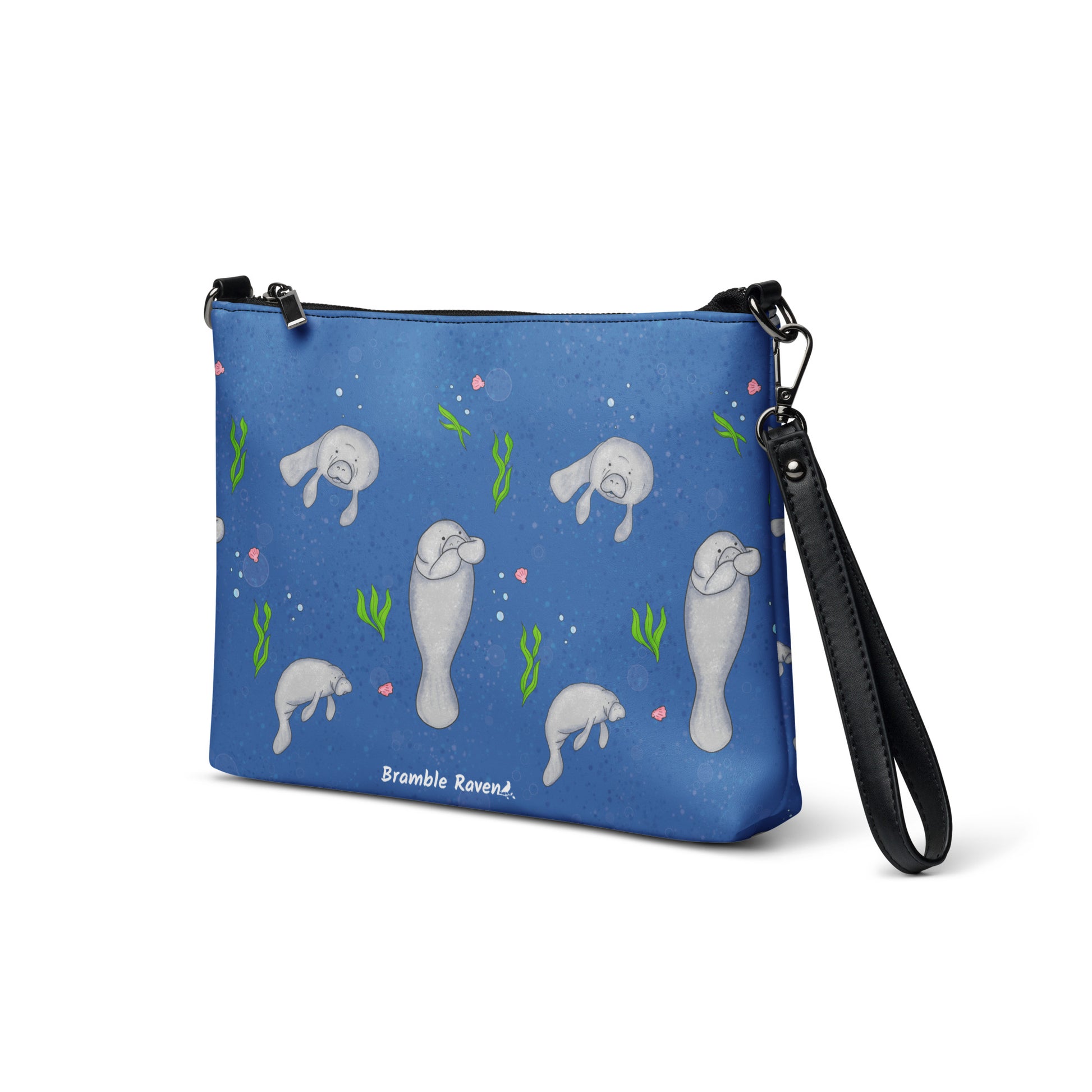 Manatee crossbody bag. Faux leather with polyester lining and dark grey hardware. Comes with adjustable removable wrist and shoulder straps. Front view of bag at an angle with manatees on a dark blue background.