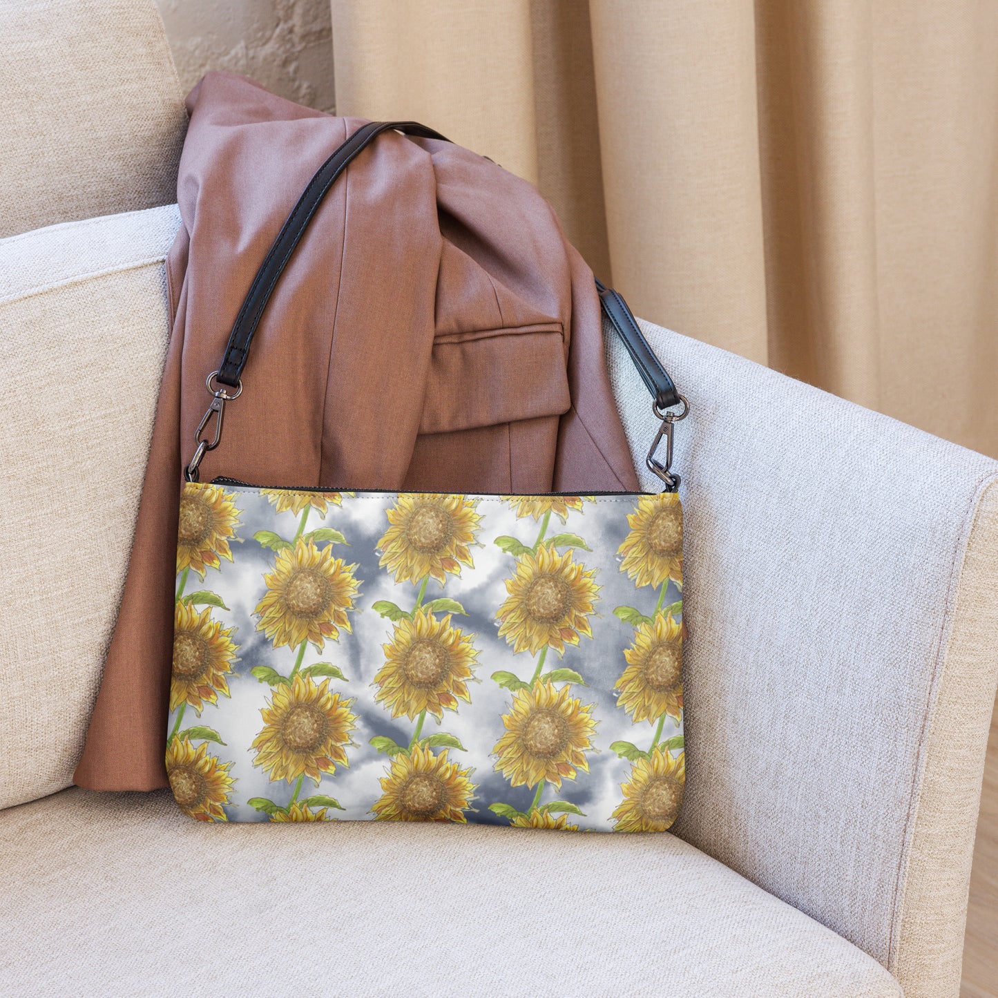 Sunflower crossbody bag. Faux leather with polyester lining and dark grey hardware. Comes with adjustable removable wrist and shoulder straps. Features patterned design of watercolor sunflowers against a cloudy background. Shown on couch by leather jacket.