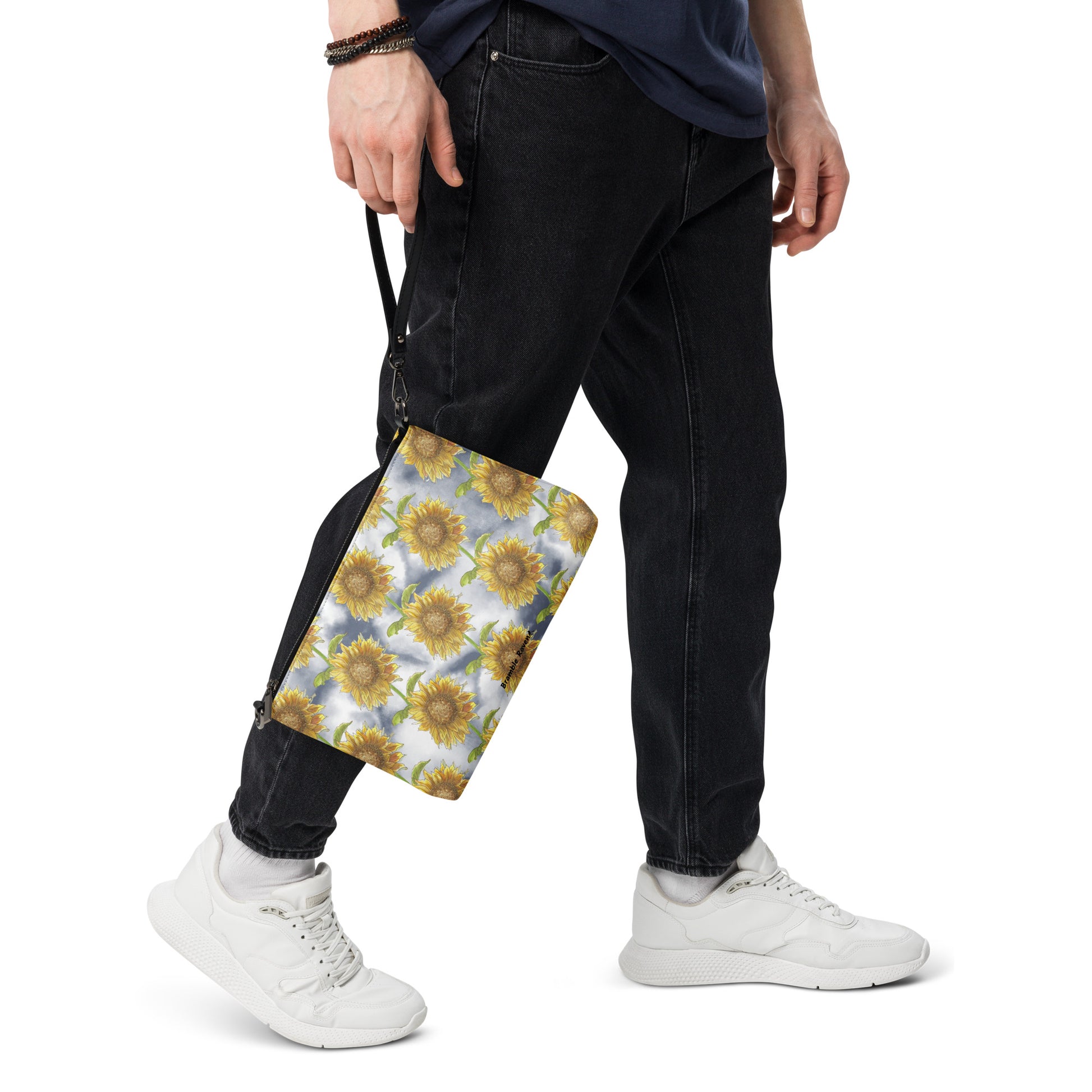 Sunflower crossbody bag. Faux leather with polyester lining and dark grey hardware. Comes with adjustable removable wrist and shoulder straps. Features patterned design of watercolor sunflowers against a cloudy background. Shown being held by wrist strap by model.