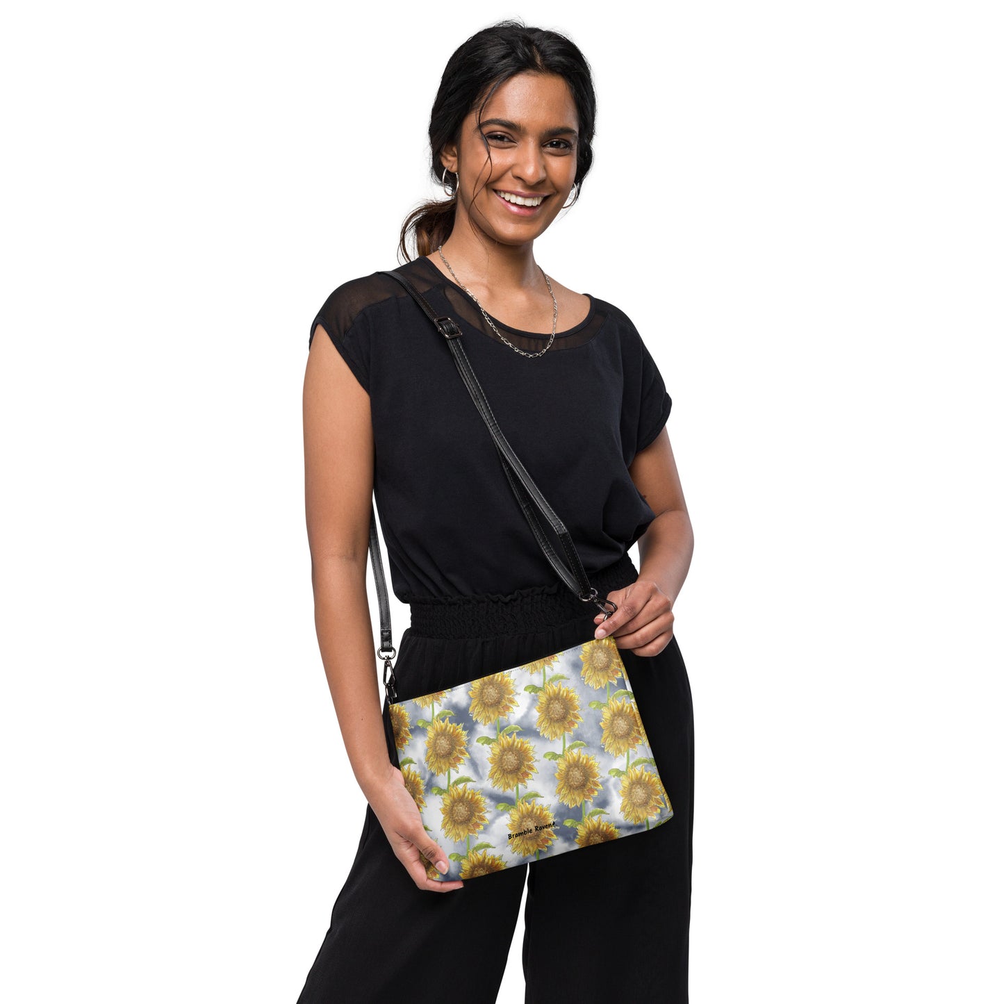 Sunflower crossbody bag. Faux leather with polyester lining and dark grey hardware. Comes with adjustable removable wrist and shoulder straps. Features patterned design of watercolor sunflowers against a cloudy background. Shown on shoulder of female model.