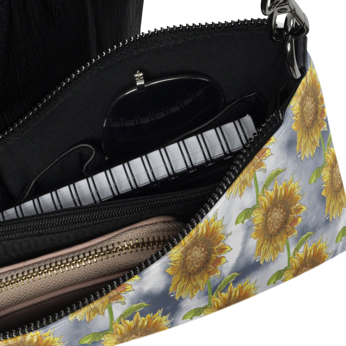 Sunflower crossbody bag. Faux leather with polyester lining and dark grey hardware. Comes with adjustable removable wrist and shoulder straps. Features patterned design of watercolor sunflowers against a cloudy background. Detail view of zipper enclosure and inside pockets. Shown with wallet, notebook, and sunglasses inside.