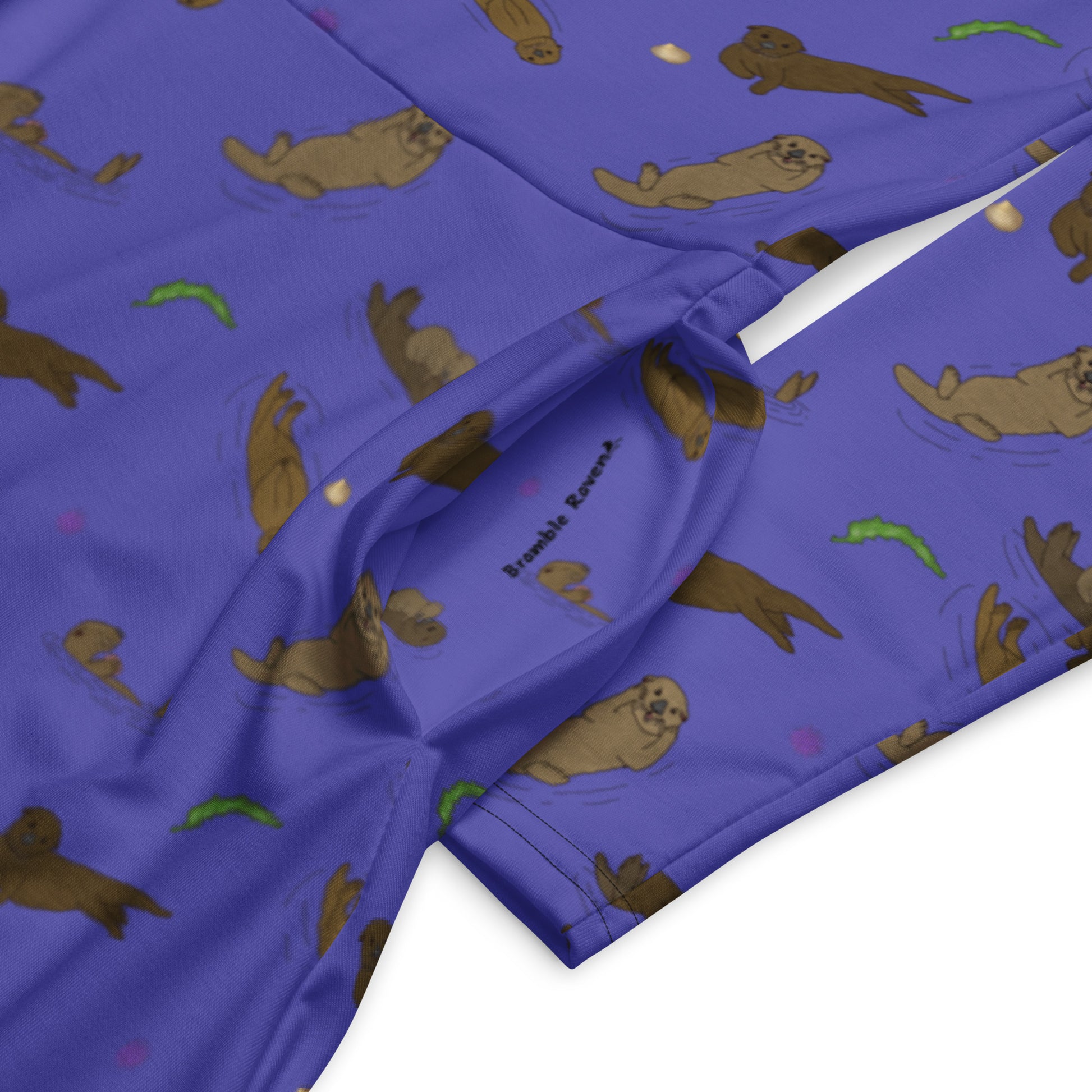 Long sleeve midi dress with fitted waist, flared bottom, and side pockets. Features a patterned design of sea otters, seashells, seaweed, and sea urchins on a purple background. Detail view of pocket with Bramble Raven logo.
