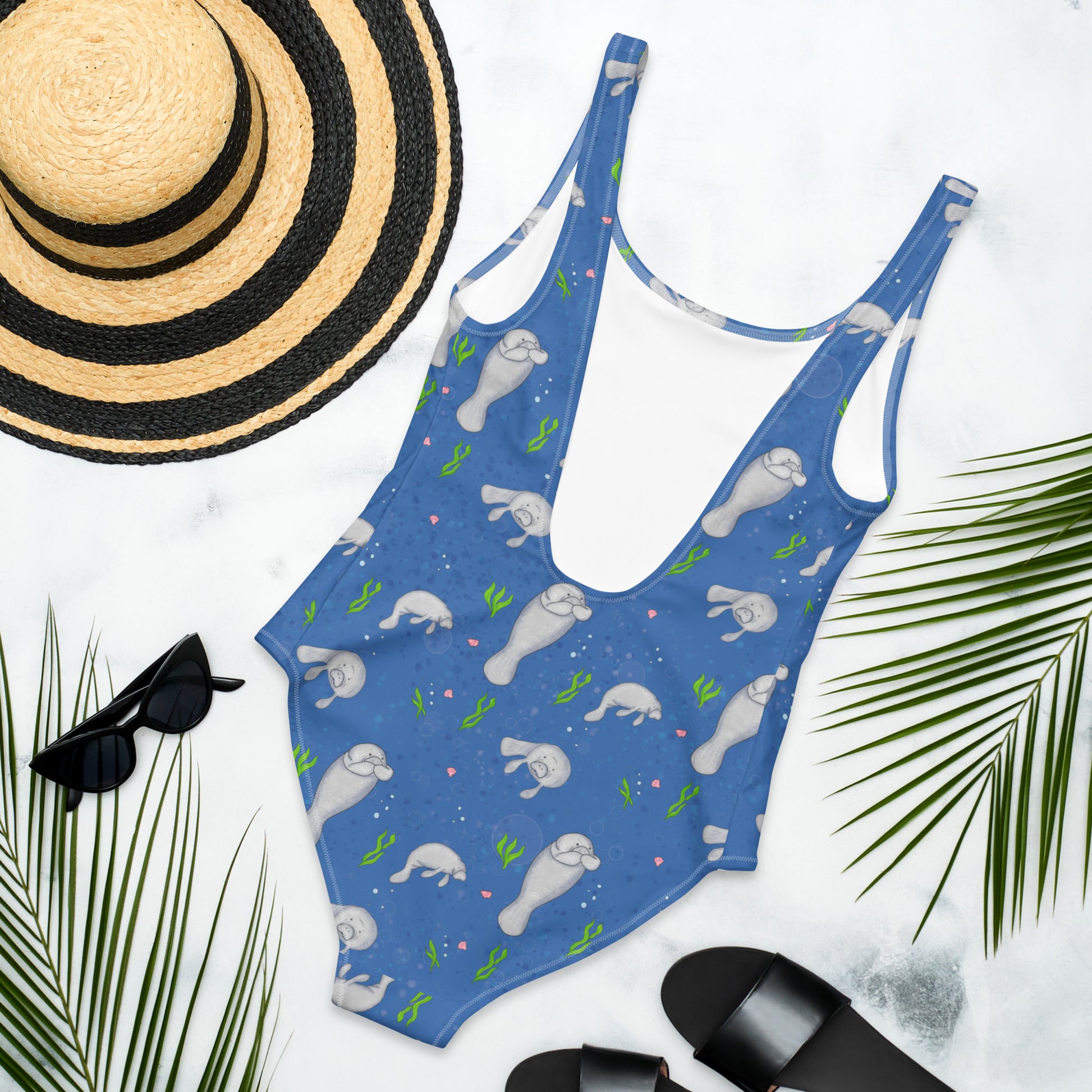 One piece swimsuit made of four-way stretch chlorine-resistant fabric. Features hand-illustrated pattern design of manatees, seaweed, shells, and bubbles on a dark blue background. Available in sizes XS to 3XL. Flat lay view of the back, by a sunhat, sunglasses, sandals and palm leaves.