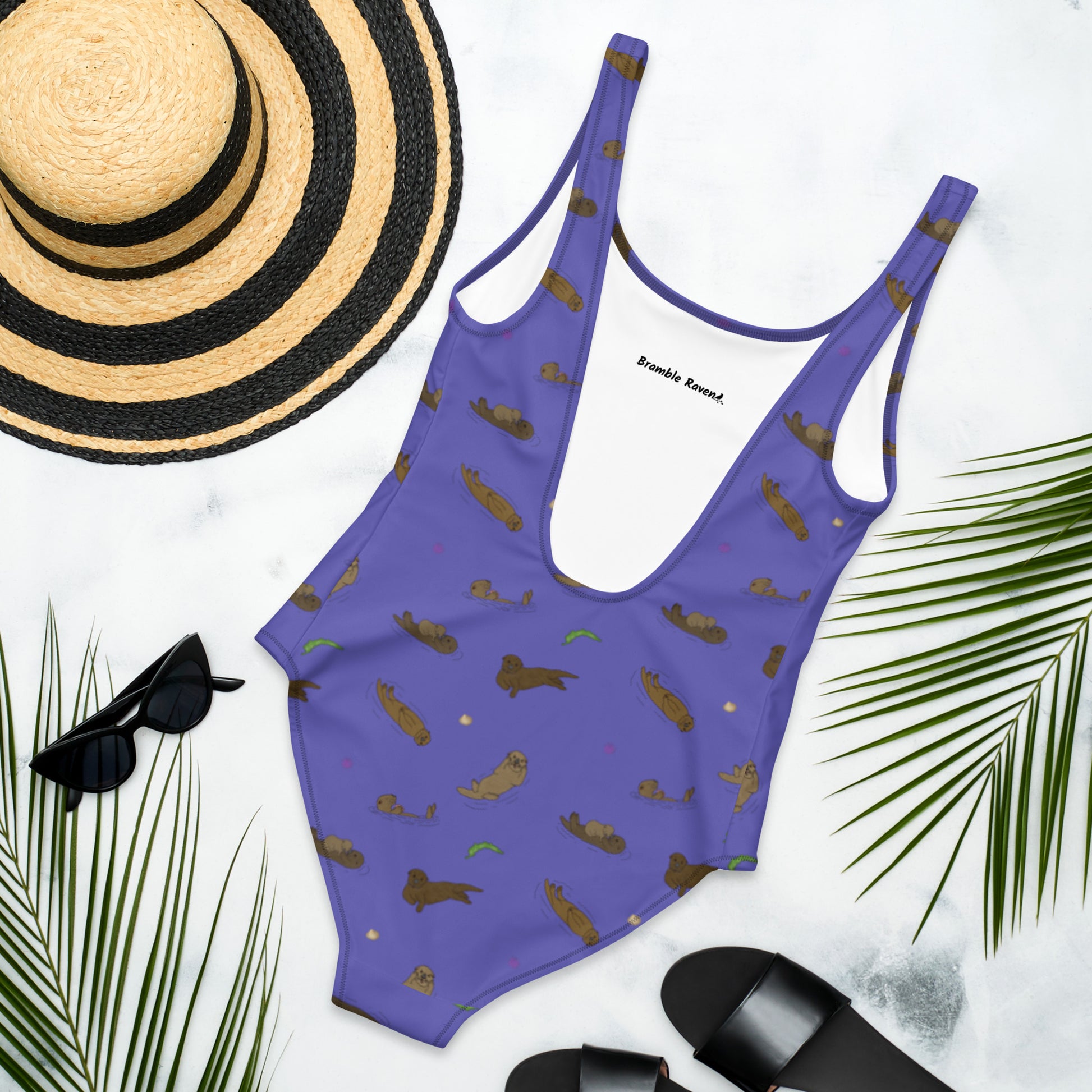 One piece swimsuit with a pattern of sea otters and ocean accents on a purple background. Image shows flat lay back view by a sunhat, sunglasses, sandals, and palm leaves.