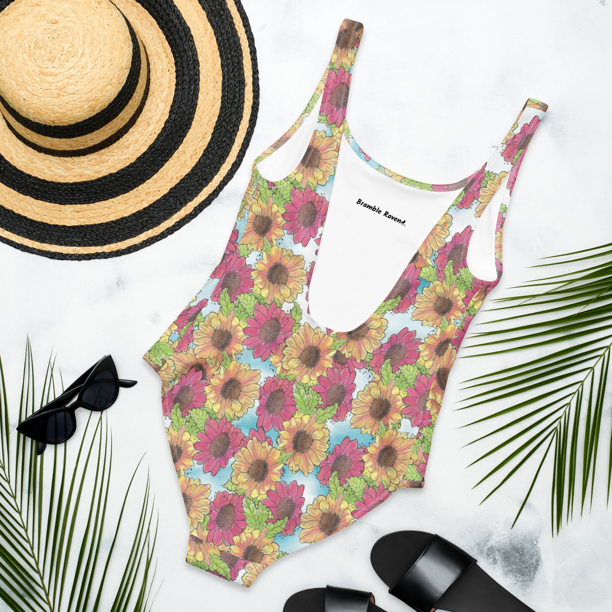 Gerber daisies one piece women's scoop back swimsuit. Yellow and red watercolor gerber daisies print. Back flat lay view by sunhat, sunglasses, and sandals. Available in XS to 3XL.