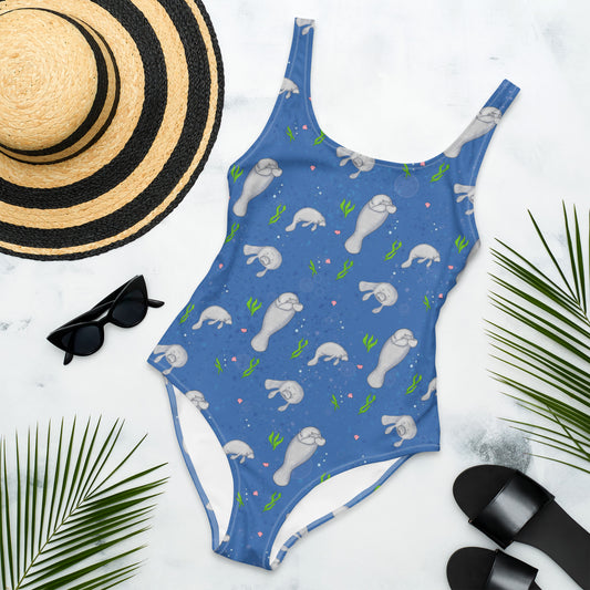 One piece swimsuit made of four-way stretch chlorine-resistant fabric. Features hand-illustrated pattern design of manatees, seaweed, shells, and bubbles on a dark blue background. Available in sizes XS to 3XL. Flat lay view of front by a sunhat, sunglasses, sandals, and palm leaves.