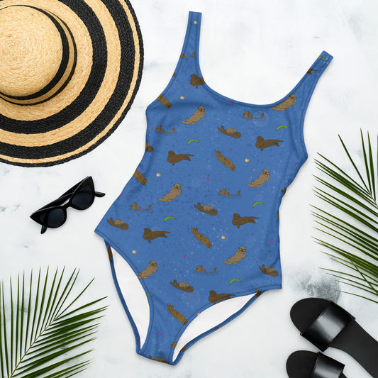 One piece swimsuit with a pattern of sea otters and ocean accents on a blue background. Image shows flat lay by a sunhat, sunglasses, sandals, and palm leaves.