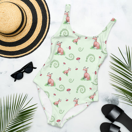 One piece swimsuit with a pattern of watercolor foxes, mushrooms and ferns on a light green background. Image shows flat lay by a sunhat, sunglasses, sandals, and palm leaves.