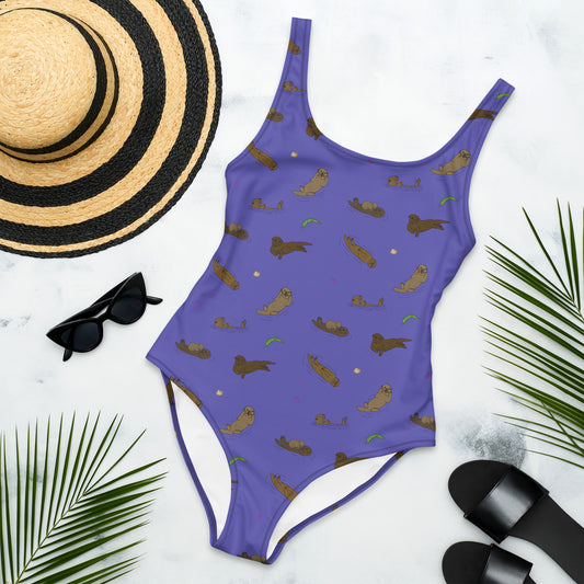One piece swimsuit with a pattern of sea otters and ocean accents on a purple background. Image shows flat lay by a sunhat, sunglasses, sandals, and palm leaves.
