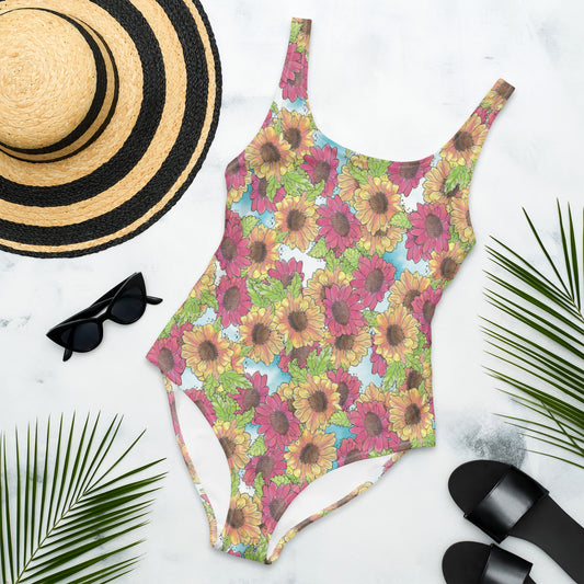 Gerber daisies one piece women's swimsuit. Yellow and red watercolor gerber daisies print. Flat lay view by sunhat, sunglasses, and sandals. Available in XS to 3XL.