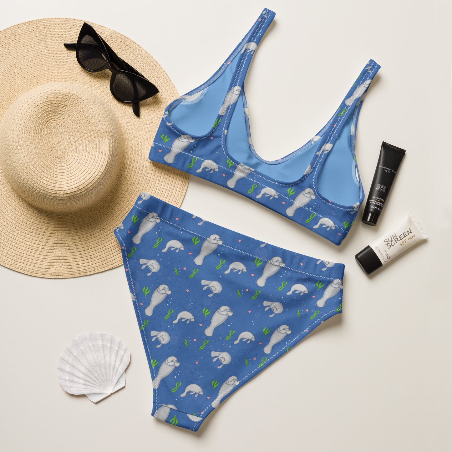 High-waisted bikini with hand-illustrated manatee pattern on a blue background. Made from recycled polyester combined with stretchable fabric. Has double layers and removable pads. Flay lay view of back, next to sunhat, sunglasses, sunscreen, and seashell.