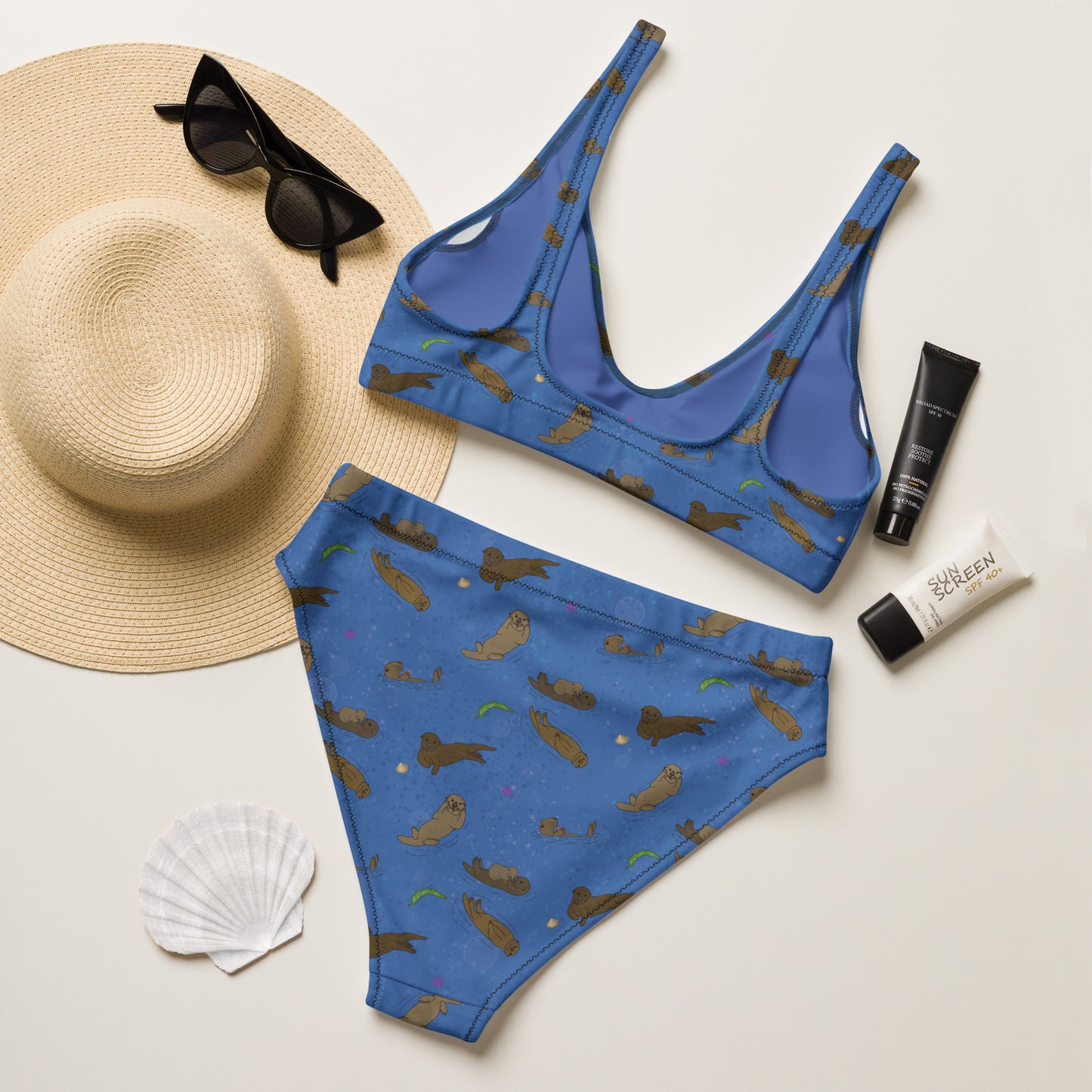 High-waisted bikini with hand-illustrated sea otters patterned on an ocean blue background. Made from recycled polyester combined with stretchable fabric. Has double layers and removable pads. Flat lay view of back shown by sunhat, sunglasses, seashell, and sunscreen.