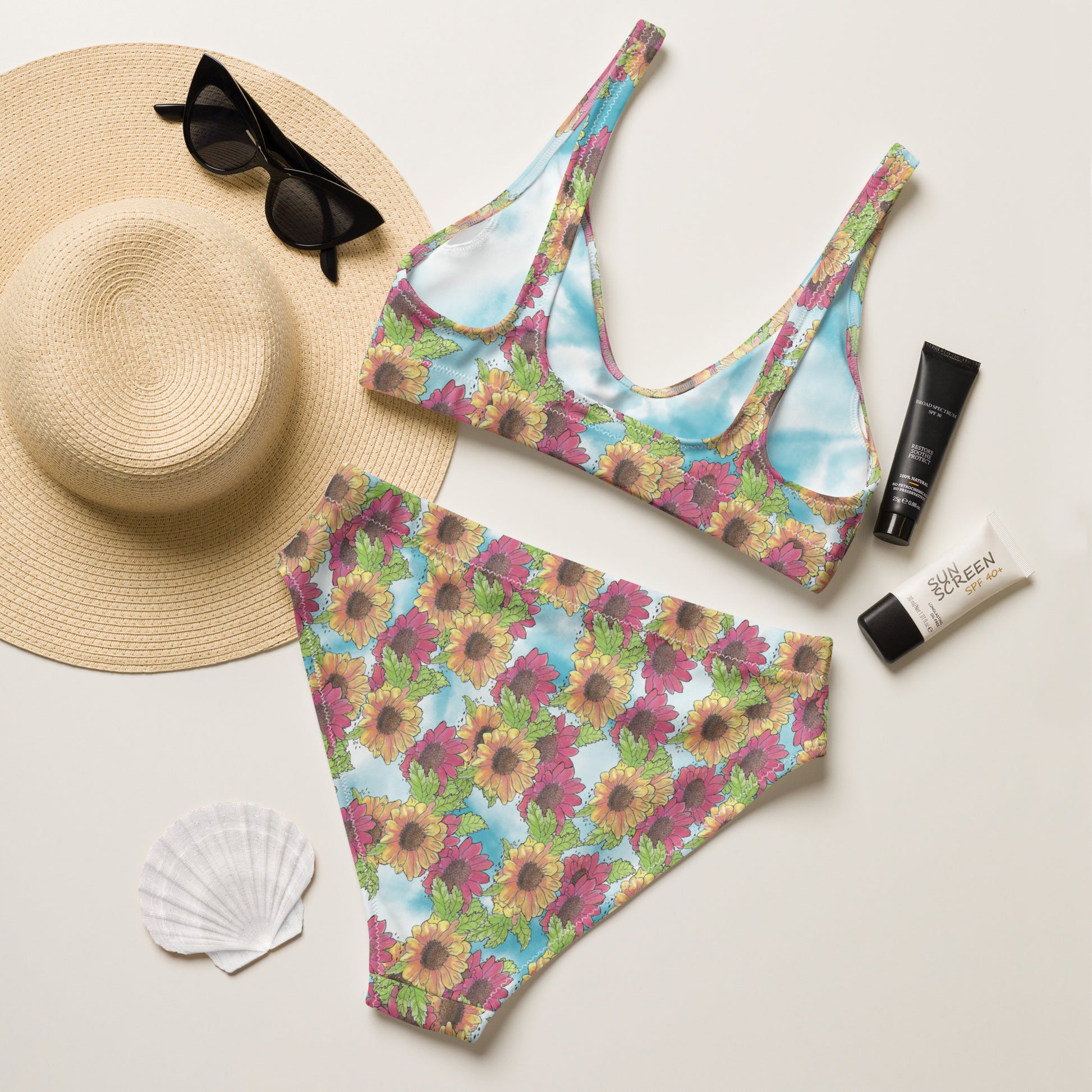 Watercolor Gerber daisy patterned high-waisted bikini. Made from recycled polyester and spandex. Features removable pads and double layers. Back flat lay view shown by sunhat, sunglasses, seashell and sunscreen.