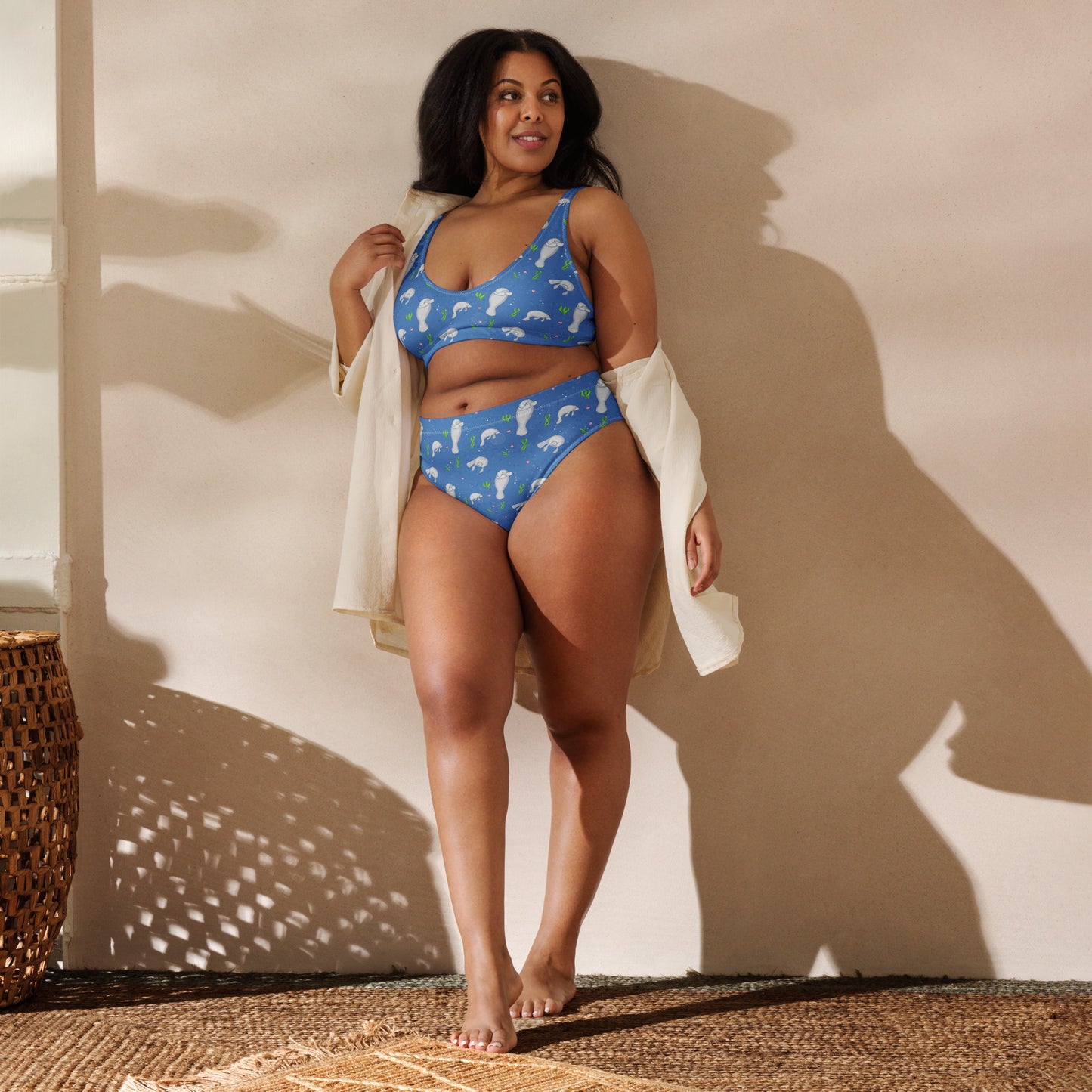 High-waisted bikini with hand-illustrated manatee pattern on a blue background. Made from recycled polyester combined with stretchable fabric. Has double layers and removable pads. Front view shown on female model leaning against a wall.