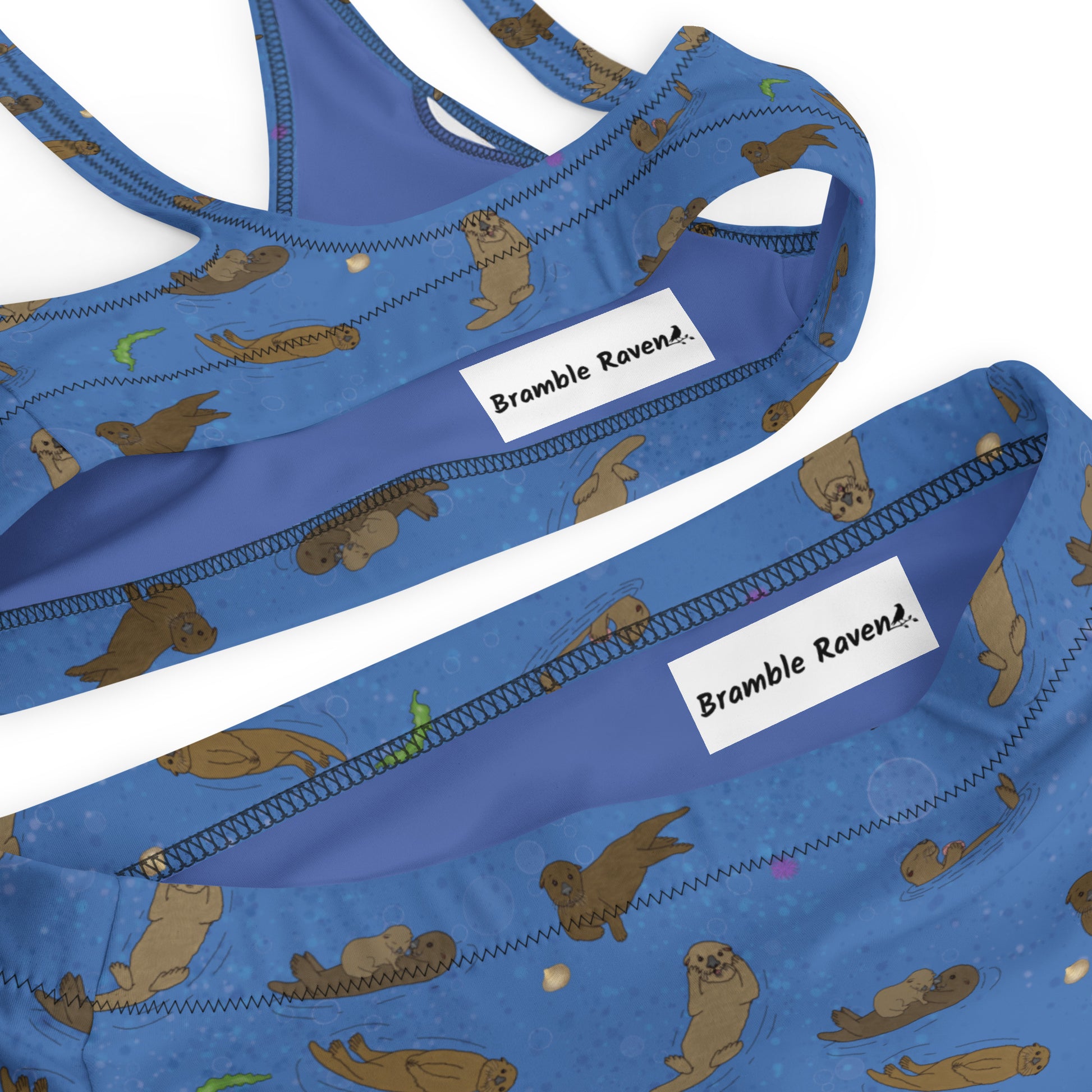 High-waisted bikini with hand-illustrated sea otters patterned on an ocean blue background. Made from recycled polyester combined with stretchable fabric. Has double layers and removable pads. Detail image of zigzag stitch and printed inner label with Bramble Raven logo.