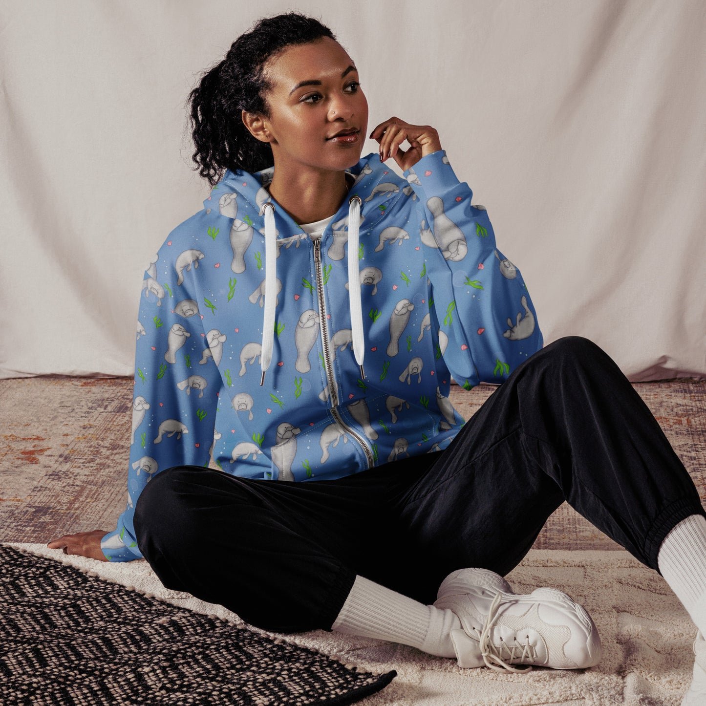 Unisex patterned manatee zip up hoodie. Made with recycled polyester. Soft cotton-feel outside fabric and fleece inside. Metal zipper, eyelets, and drawcord tips. Shown on female model sitting on carpet.
