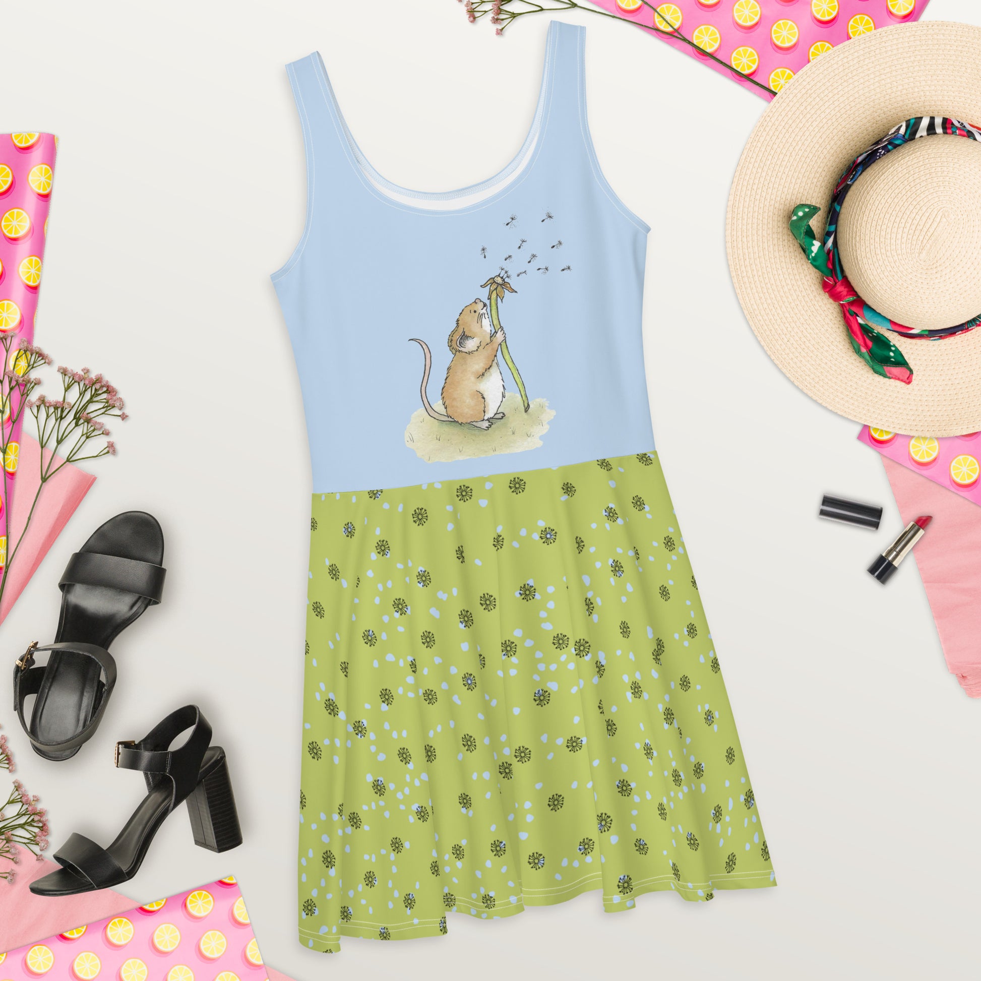 Sleeveless skater dress featuring Dandelion Wish design of a mouse making a wish on a dandelion fluff on a blue top. The mid-thigh length flared green skirt features flowers and dot details. Also has an elastic waistline. Flat lay view on floor by sunhat, black high heels, lipstick, pink polka dot wrapping paper, and flowers.