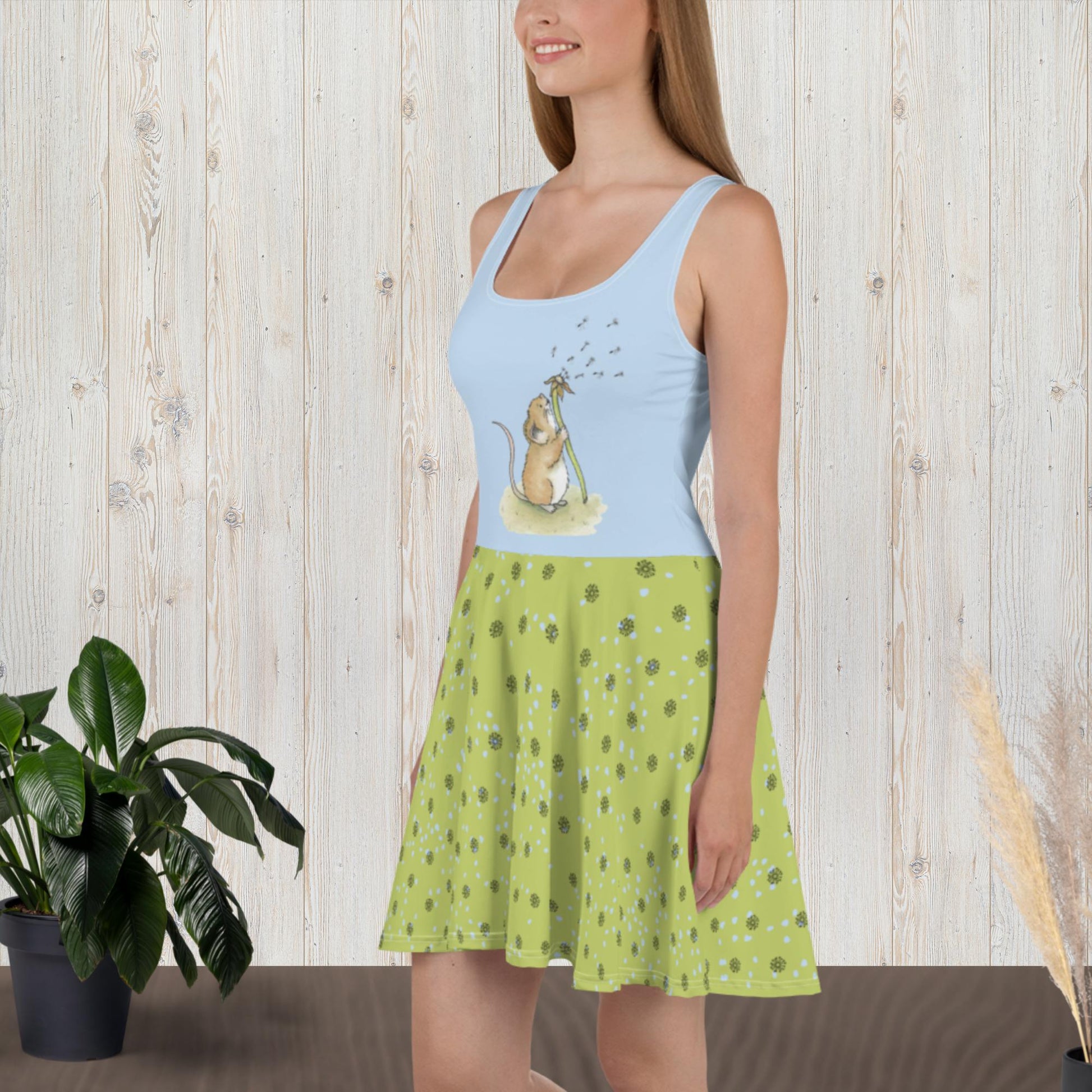 Sleeveless skater dress featuring Dandelion Wish design of a mouse making a wish on a dandelion fluff on a blue top. The mid-thigh length flared green skirt features flowers and dot details. Also has an elastic waistline. Shown on female model facing left.
