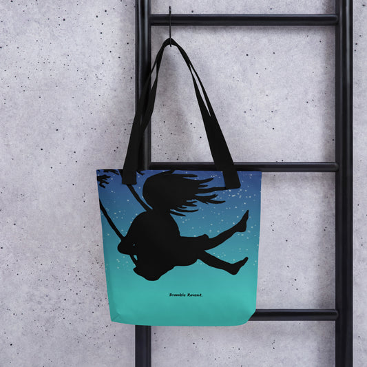 15 by 15 inch sturdy night swing tote bag. Has black handles and features a girl in a tree swing against the backdrop of a starry summer night sky. Shown hanging on a black ladder.