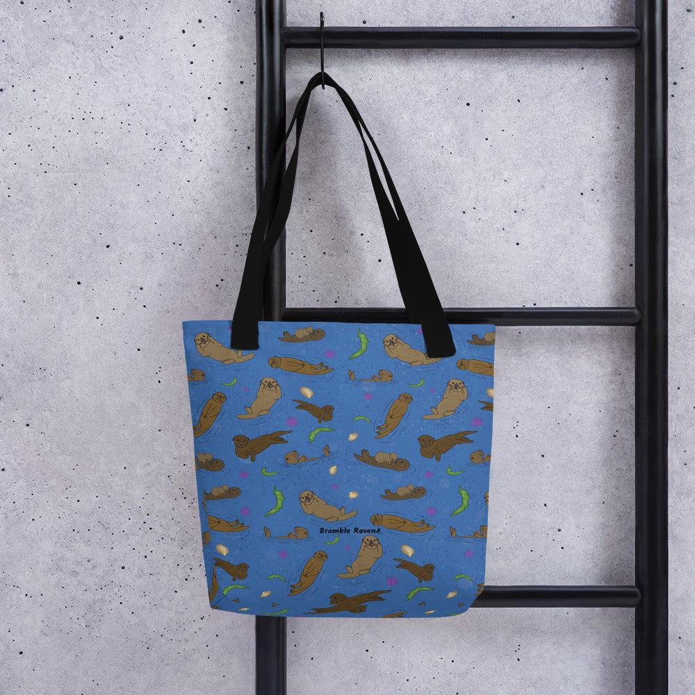 15 by 15 inch polyester tote bag with dual black cotton bull denim handles. It has a patterned design of sea otters, seashells, seaweed and sea urchins on a blue background. Shown hanging from a black ladder.