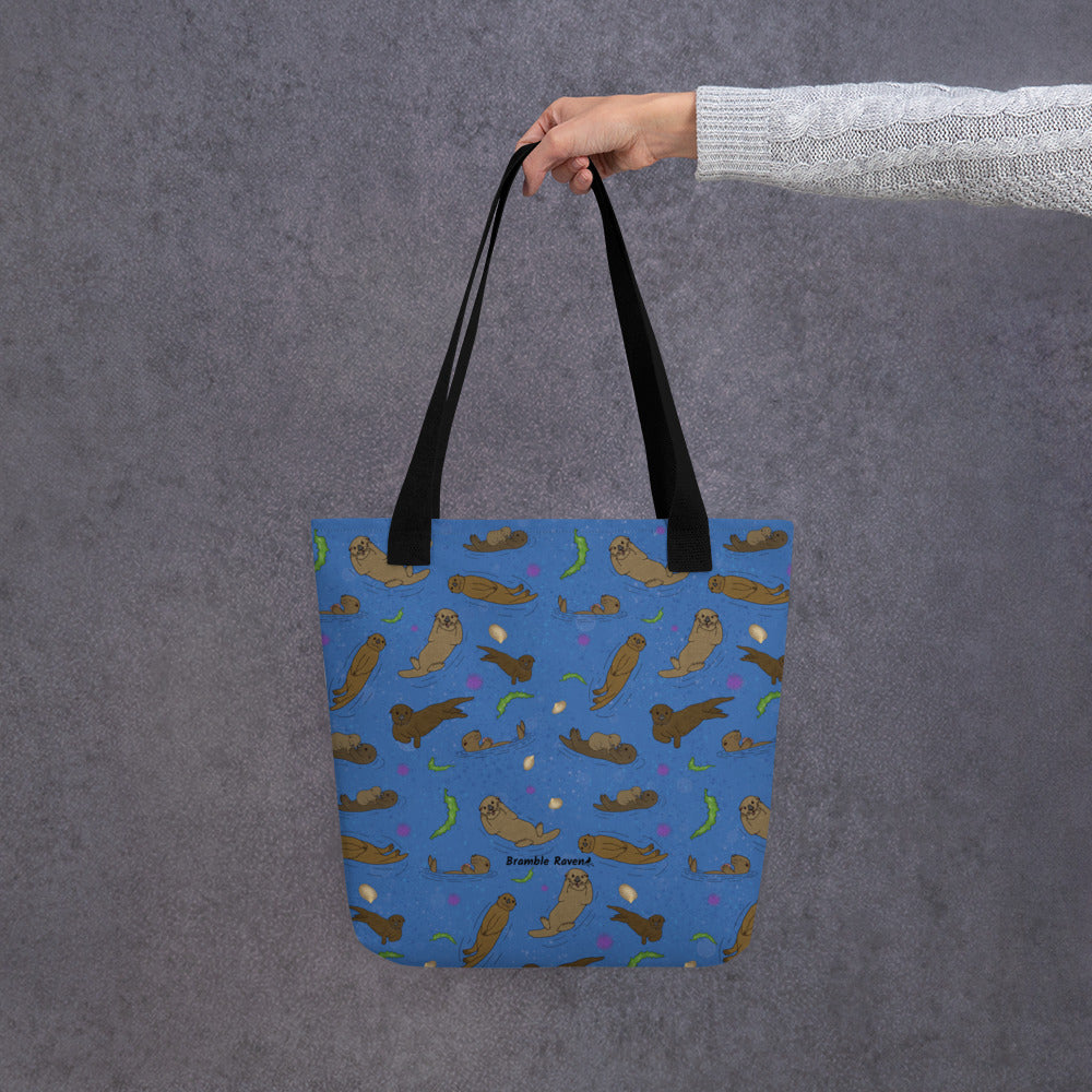 15 by 15 inch polyester tote bag with dual black cotton bull denim handles. It has a patterned design of sea otters, seashells, seaweed and sea urchins on a blue background. Shown hanging from a model's hand.