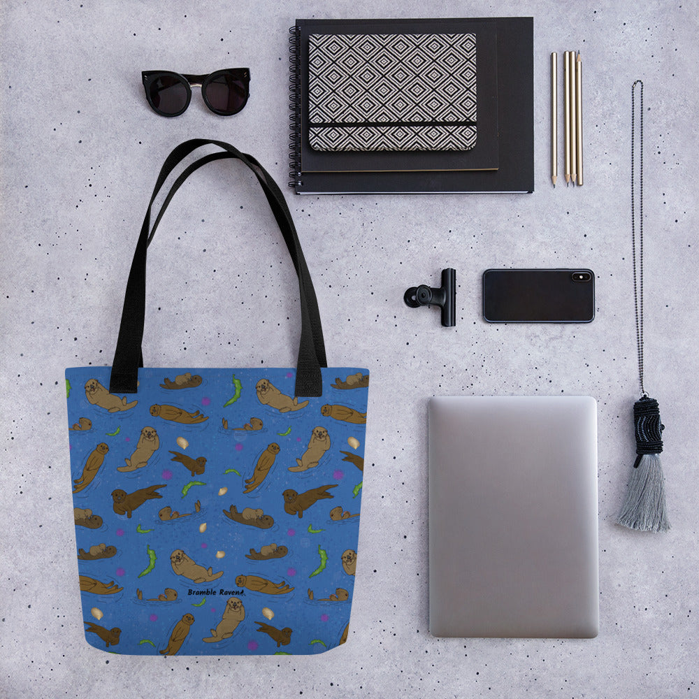 15 by 15 inch polyester tote bag with dual black cotton bull denim handles. It has a patterned design of sea otters, seashells, seaweed and sea urchins on a blue background. Flay lay by a notebook, sunglasses, pencils, clip, necklace, phone, and tablet.