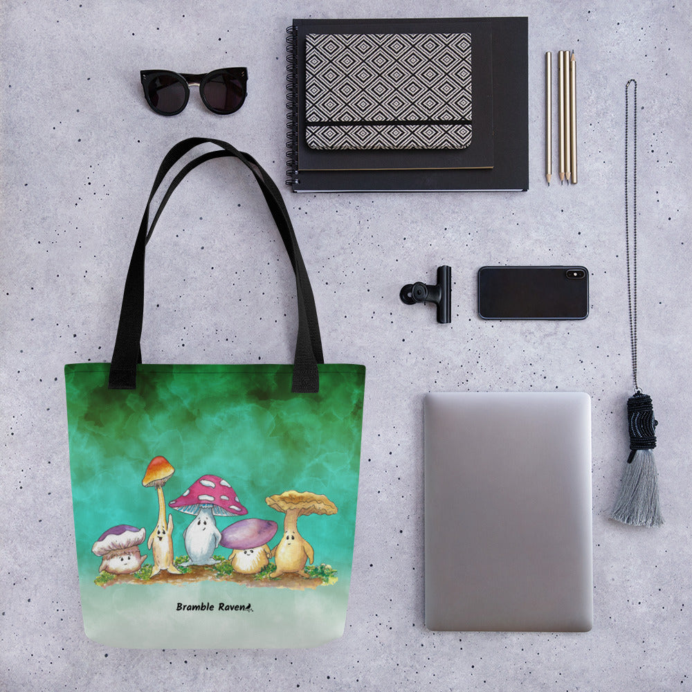 15 by 15 inch sturdy tote bag. Features Mushy and his whimsical mushroom friends on a green gradient background. Made of spun polyester with black cotton bull denim handles. Shown on table by sunglasses, notebook, tablet, phone, and keychain.