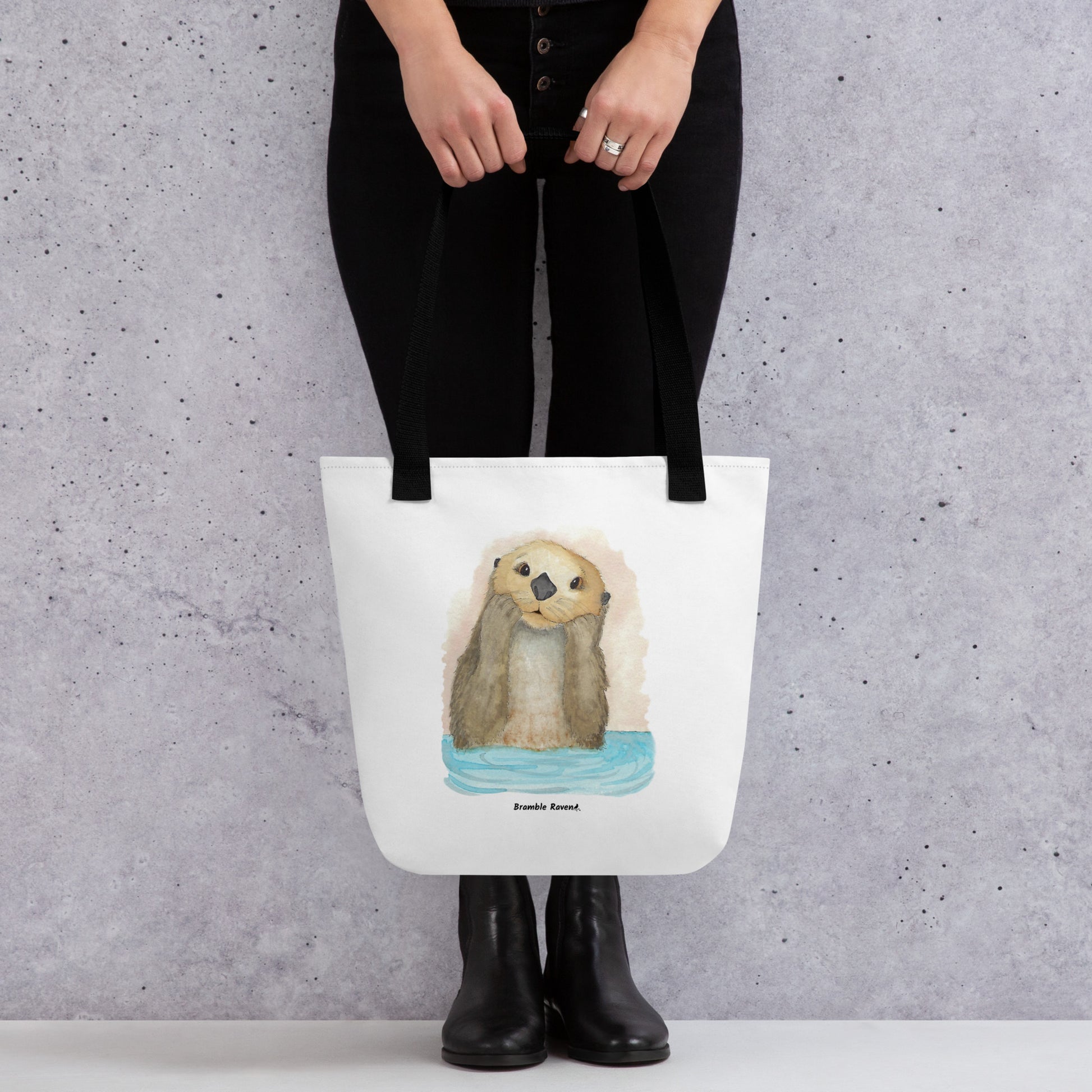 Otter Amazement sturdy tote bag. Measures 15" by 15" and can hold up to 44 lbs. Features watercolor sea otter painting printed on front and back. Has black handles. Shown hanging from model's hands.