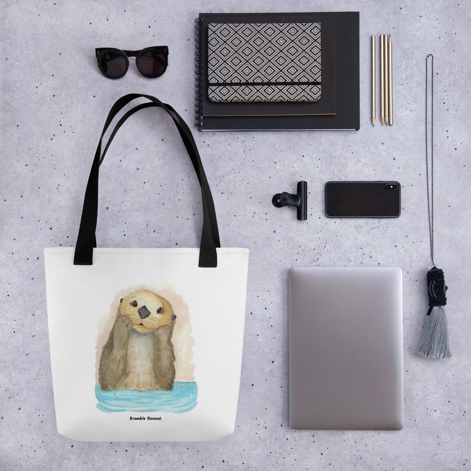 Otter Amazement sturdy tote bag. Measures 15" by 15" and can hold up to 44 lbs. Features watercolor sea otter painting printed on front and back. Has black handles. Shown by a notebook, sunglasses, phone, tablet, and keychain.