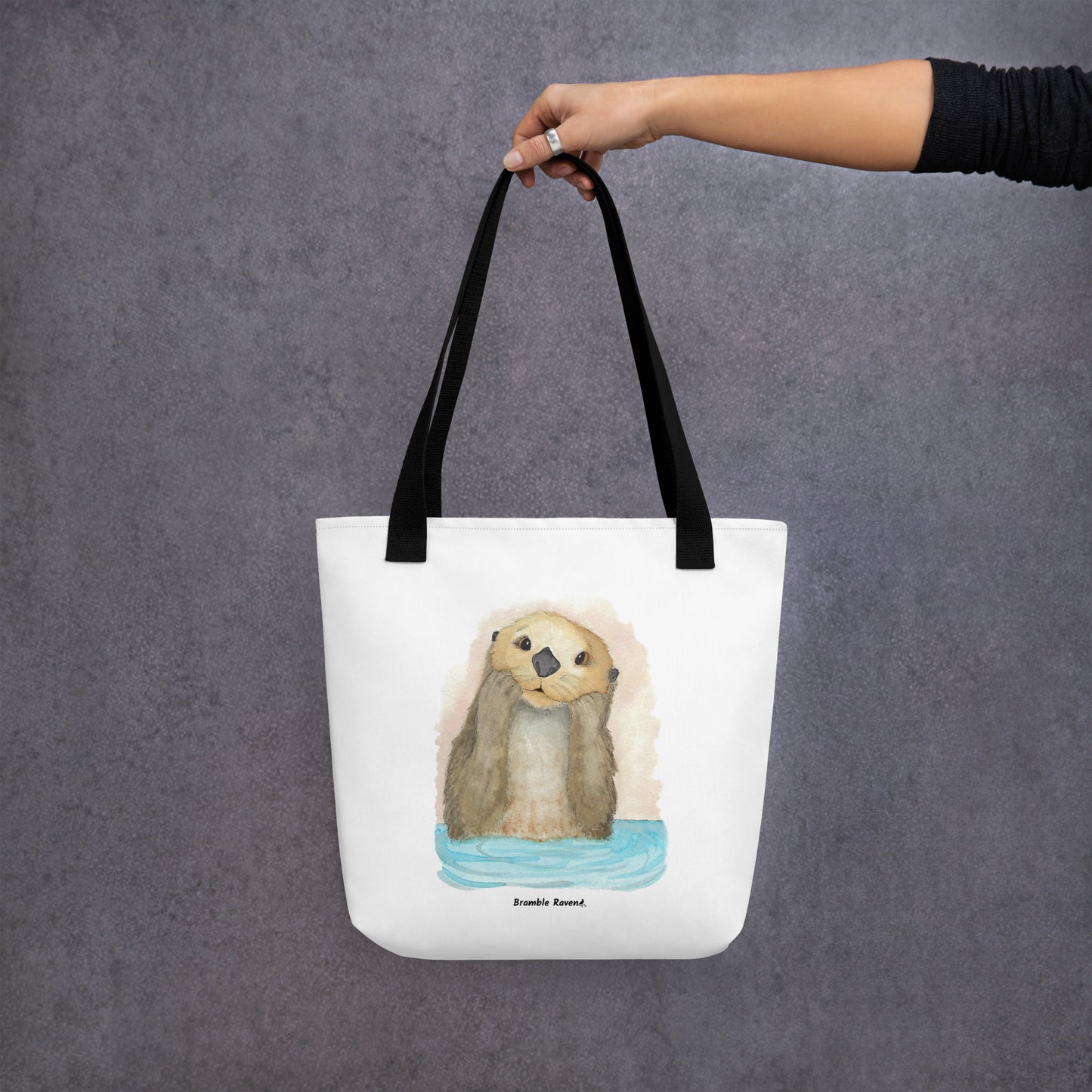 Otter Amazement sturdy tote bag. Measures 15" by 15" and can hold up to 44 lbs. Features watercolor sea otter painting printed on front and back. Has black handles. Shown hanging from a model's hand.