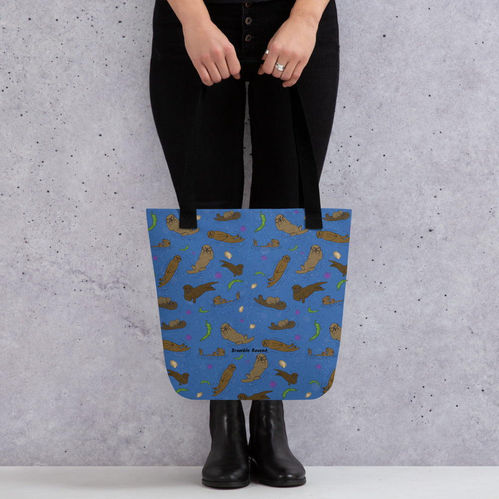 15 by 15 inch polyester tote bag with dual black cotton bull denim handles. It has a patterned design of sea otters, seashells, seaweed and sea urchins on a blue background. Shown being held in model's hands.