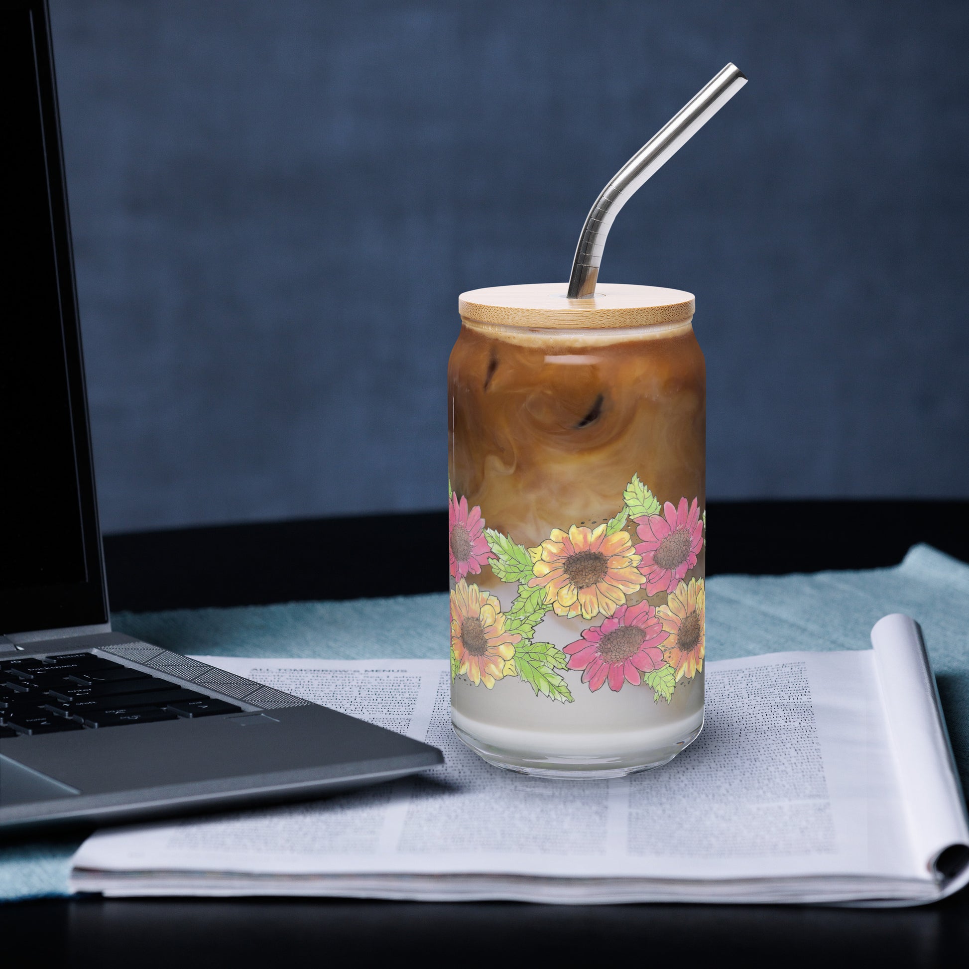 16 ounce can-shaped glass. Features wraparound print of watercolor Gerber daisies. Shown with bamboo lid and stainless steel straw, filled with iced coffee and sitting by a laptop.